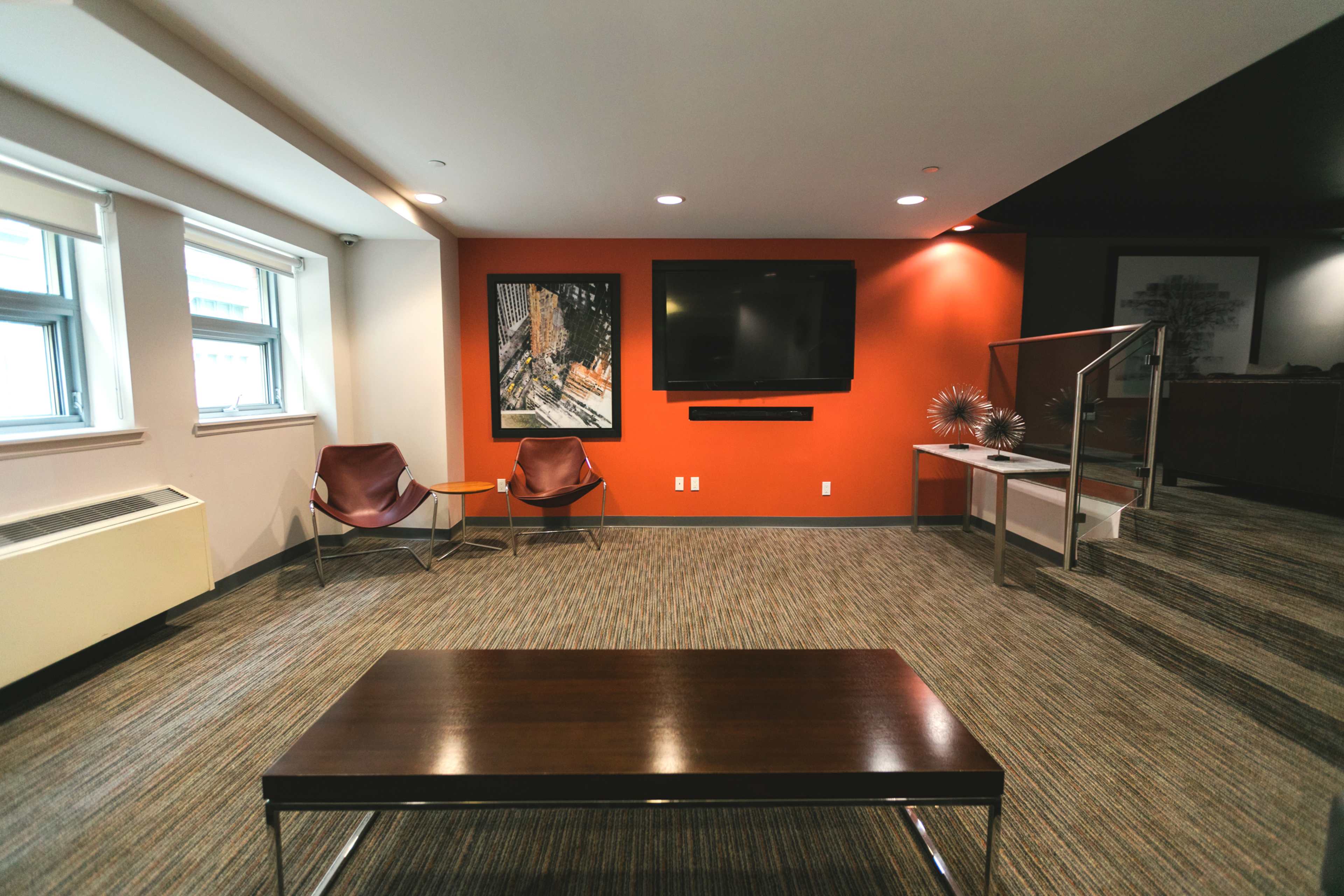 The image shows a modern living space with two chairs, a wooden coffee table, a large wall-mounted television, and artwork on the walls.