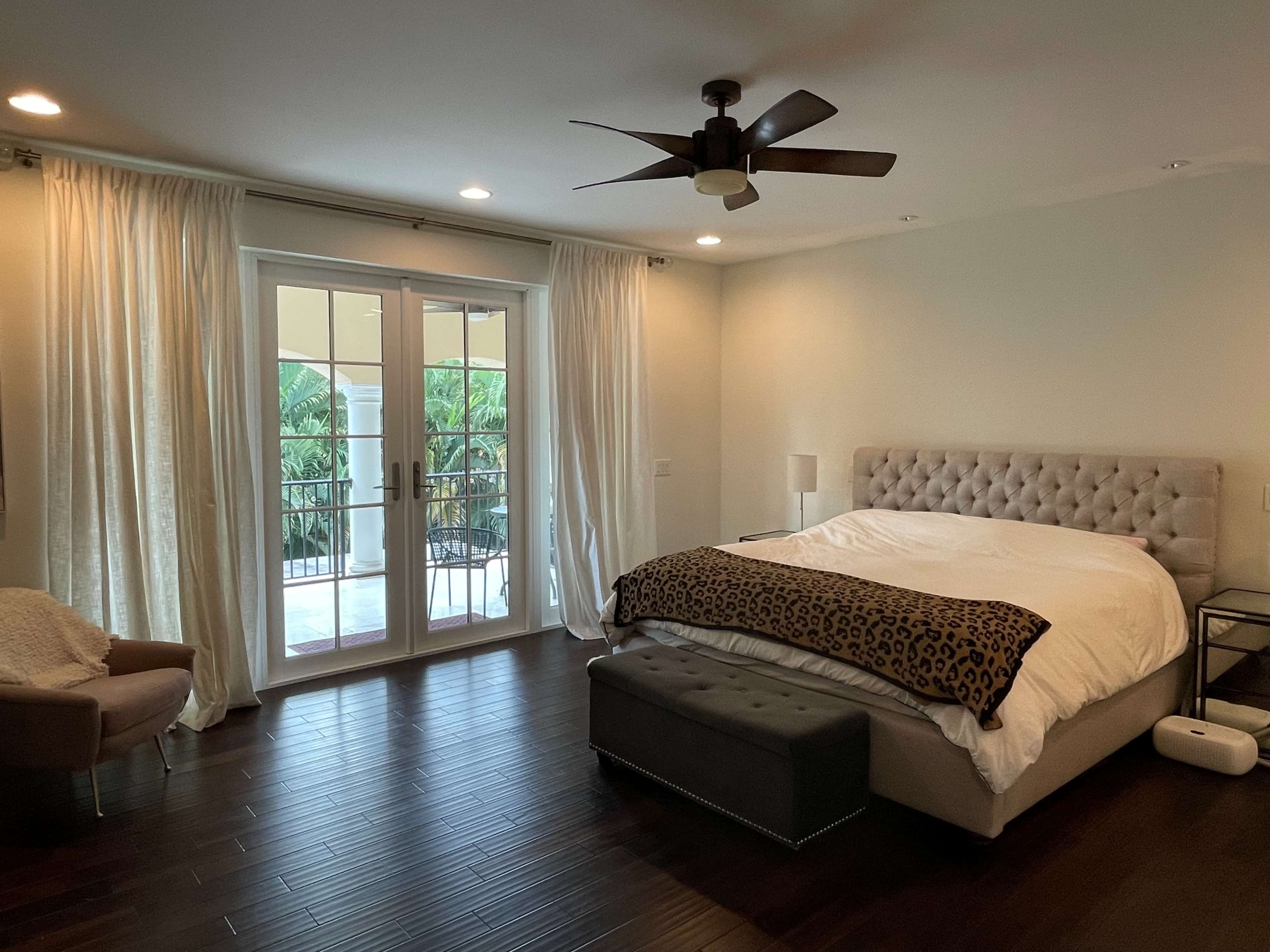 A modern bedroom features a large, tufted bed, a bench at the foot, and double French doors leading to a balcony surrounded by greenery.