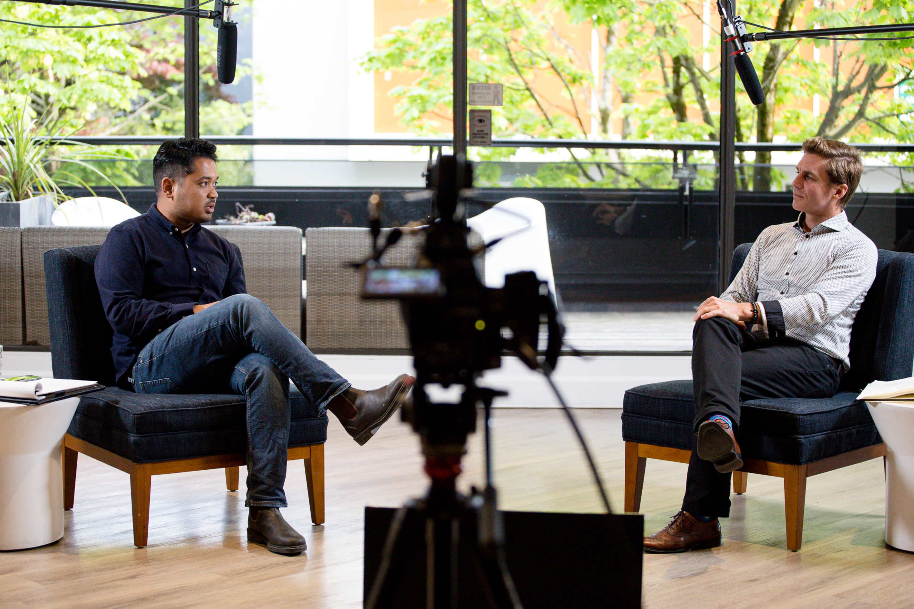 Two men are seated in a brightly lit room with large windows, while a camera pointing at them captures the conversation.