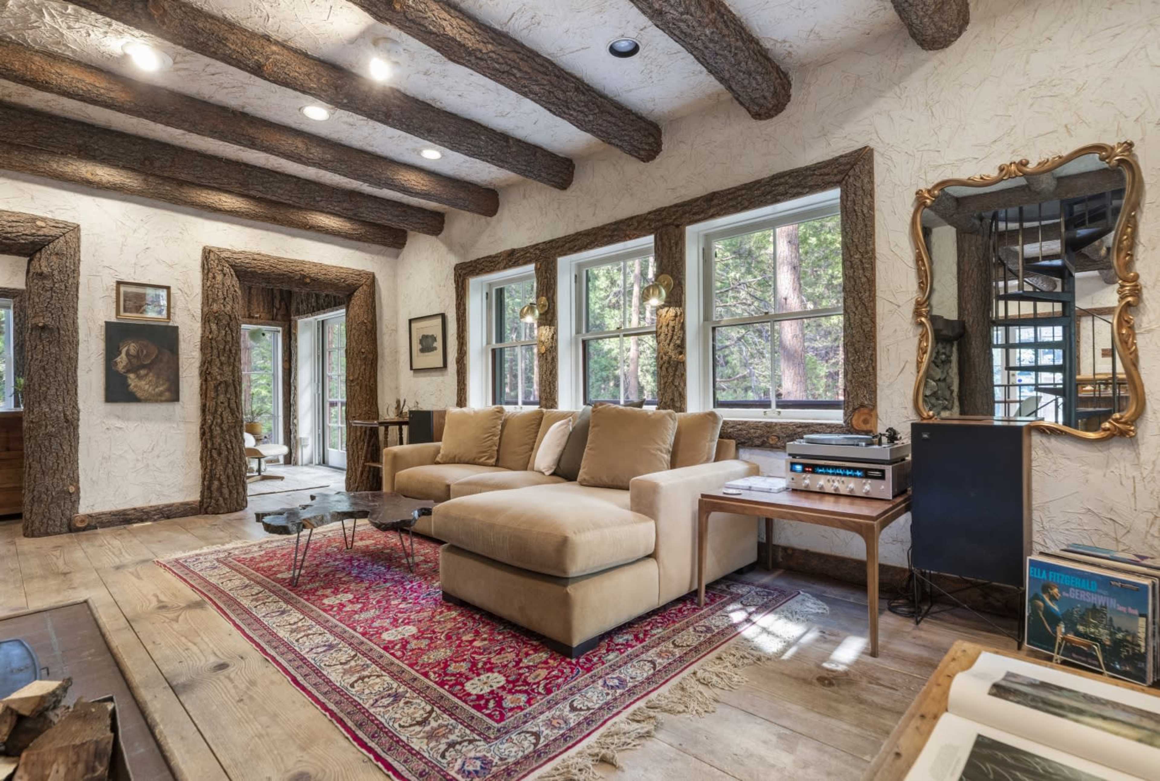The living room features a beige sectional sofa, wooden beams along the ceiling, and large windows overlooking a forested area.
