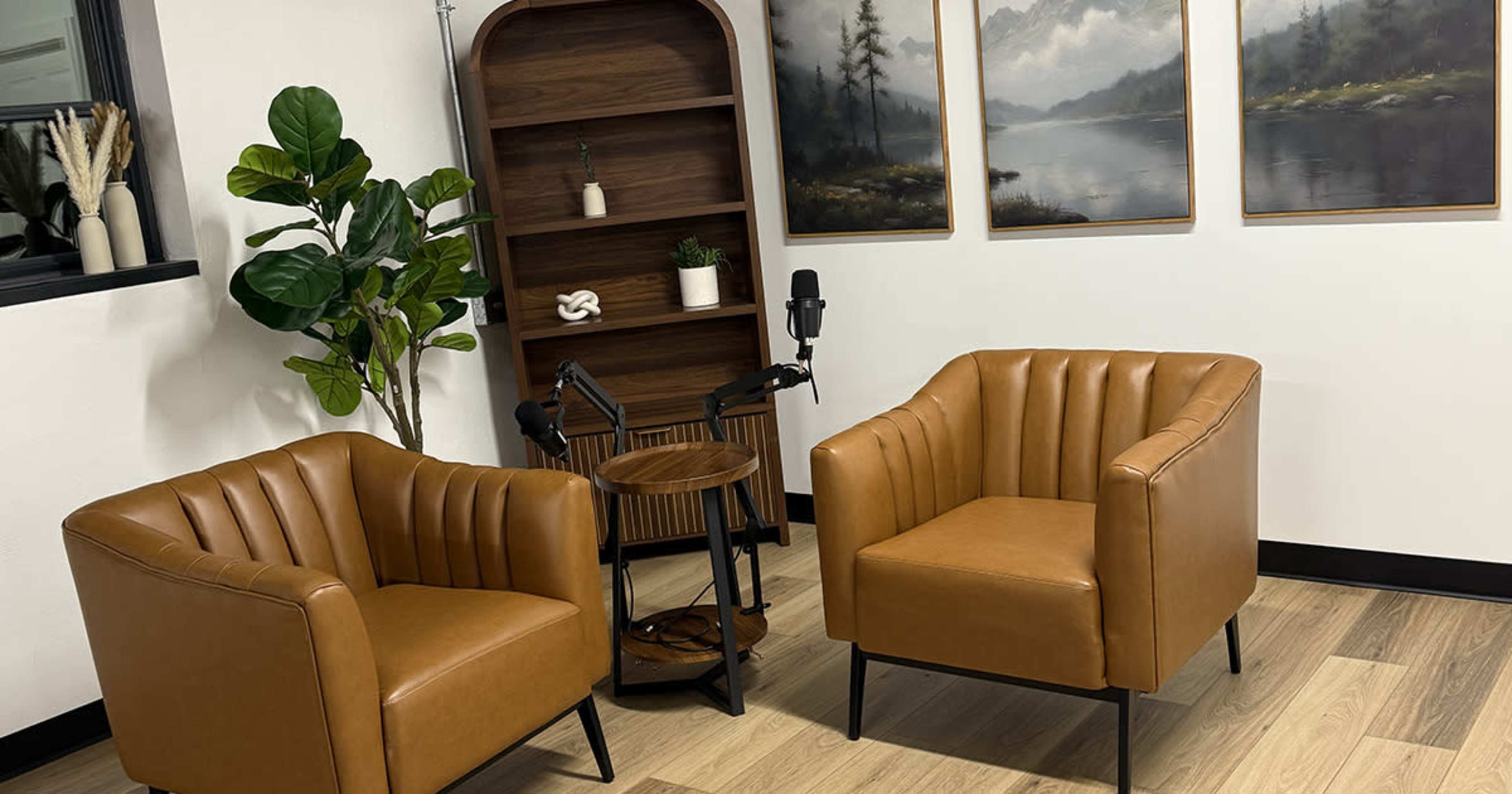 The image shows a cozy sitting area with two brown, tufted chairs facing a small table, alongside a bookcase and framed landscape paintings on the wall.