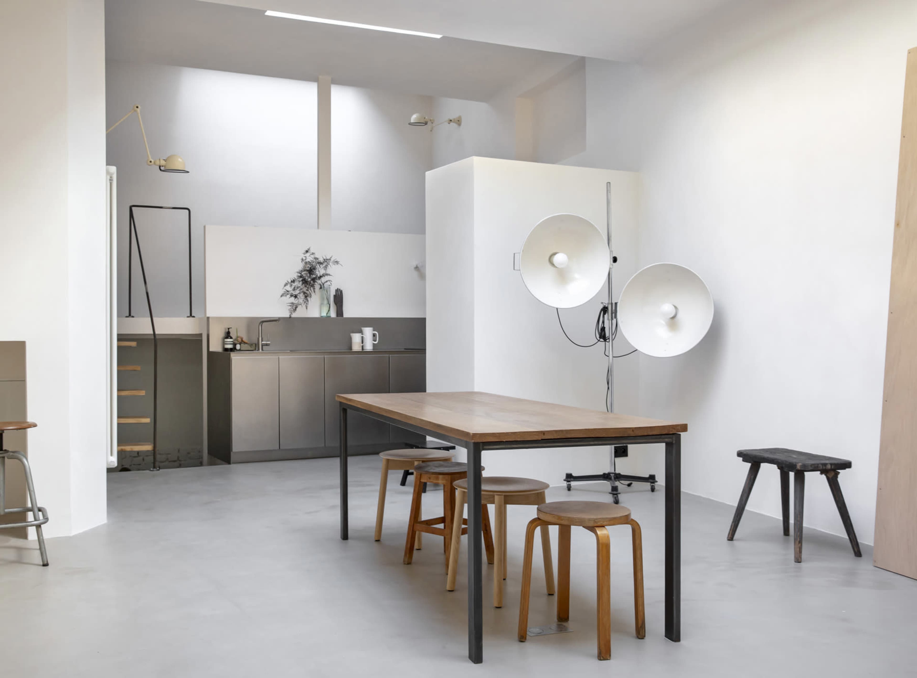 The image shows a minimalist kitchen and dining area with a wooden table surrounded by stools, and large light fixtures mounted on a wall.