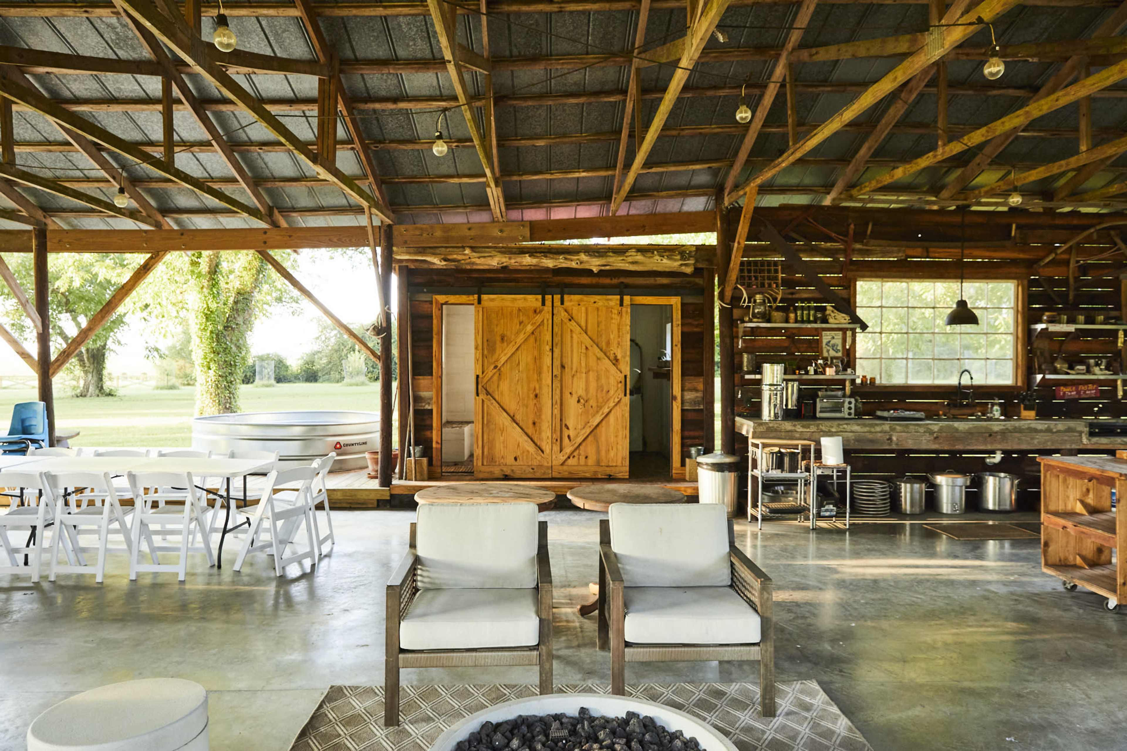 The image shows a spacious, rustic interior of a barn-like structure with seating and a kitchen area, featuring large wooden doors and large windows overlooking a grassy field.