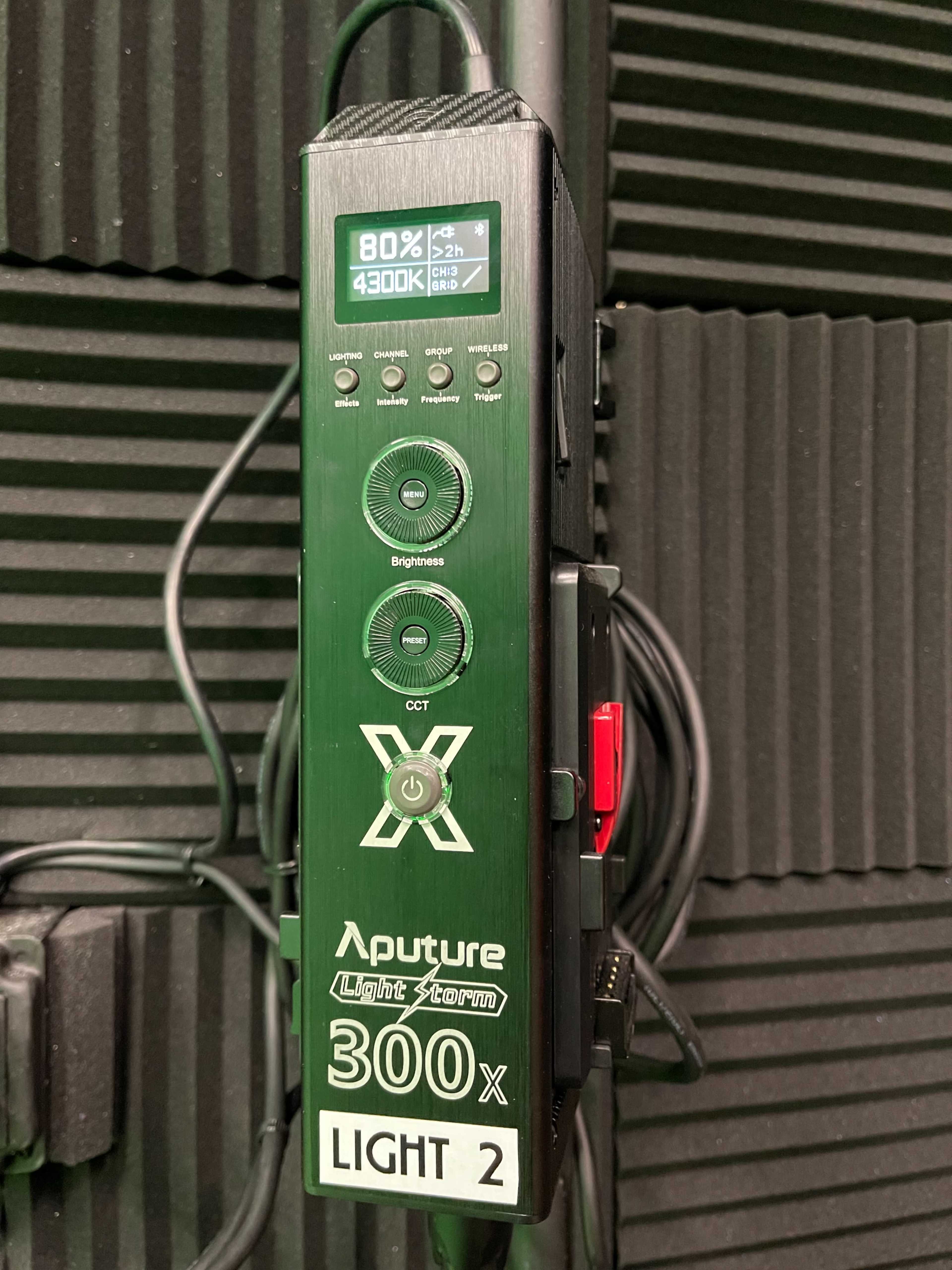 A control panel for an Aputure 300x light, featuring buttons for brightness and color temperature adjustments, mounted on a wall with soundproofing panels.