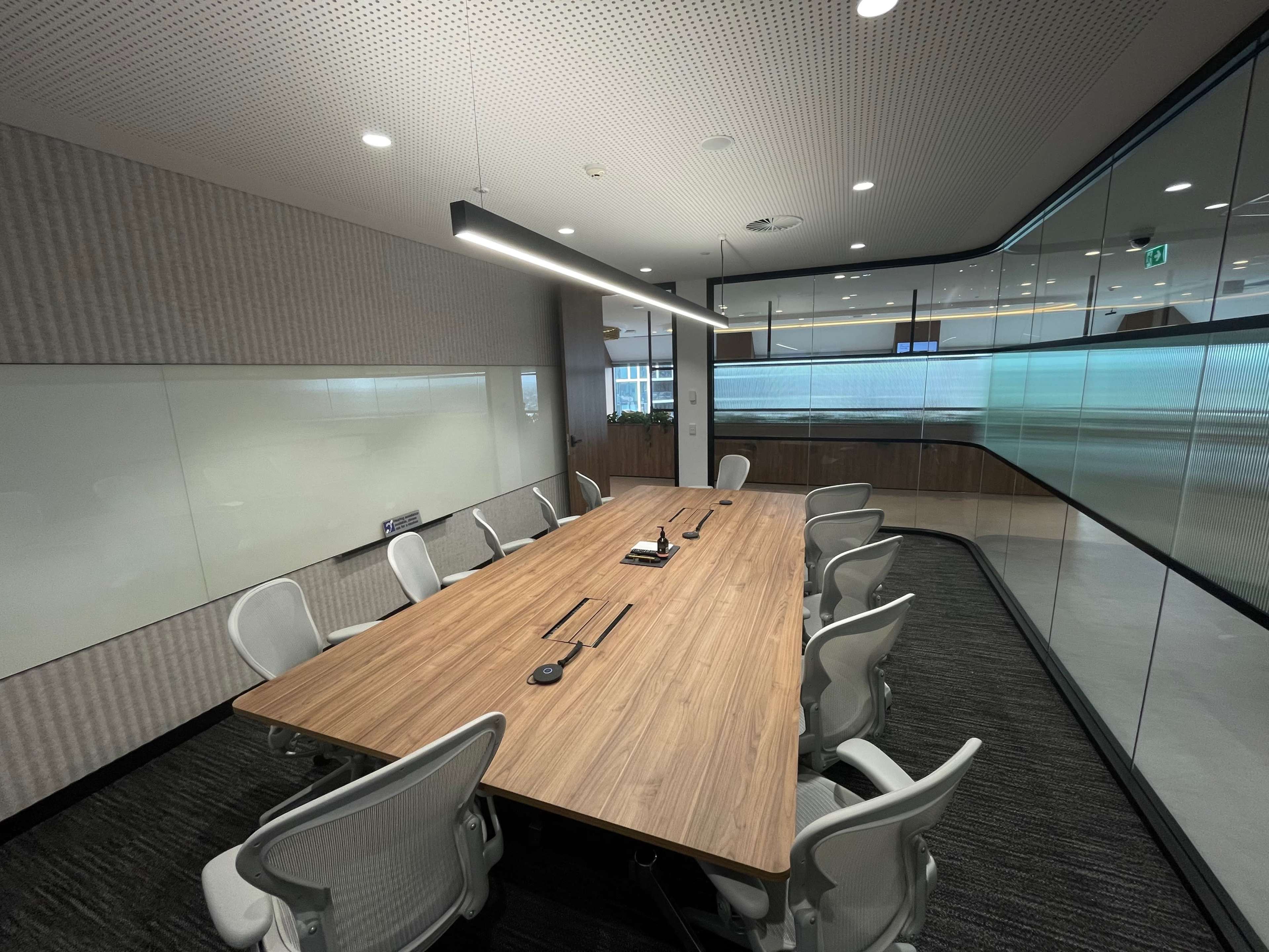 A modern conference room features a large wooden table surrounded by white ergonomic chairs, with glass walls and a whiteboard.
