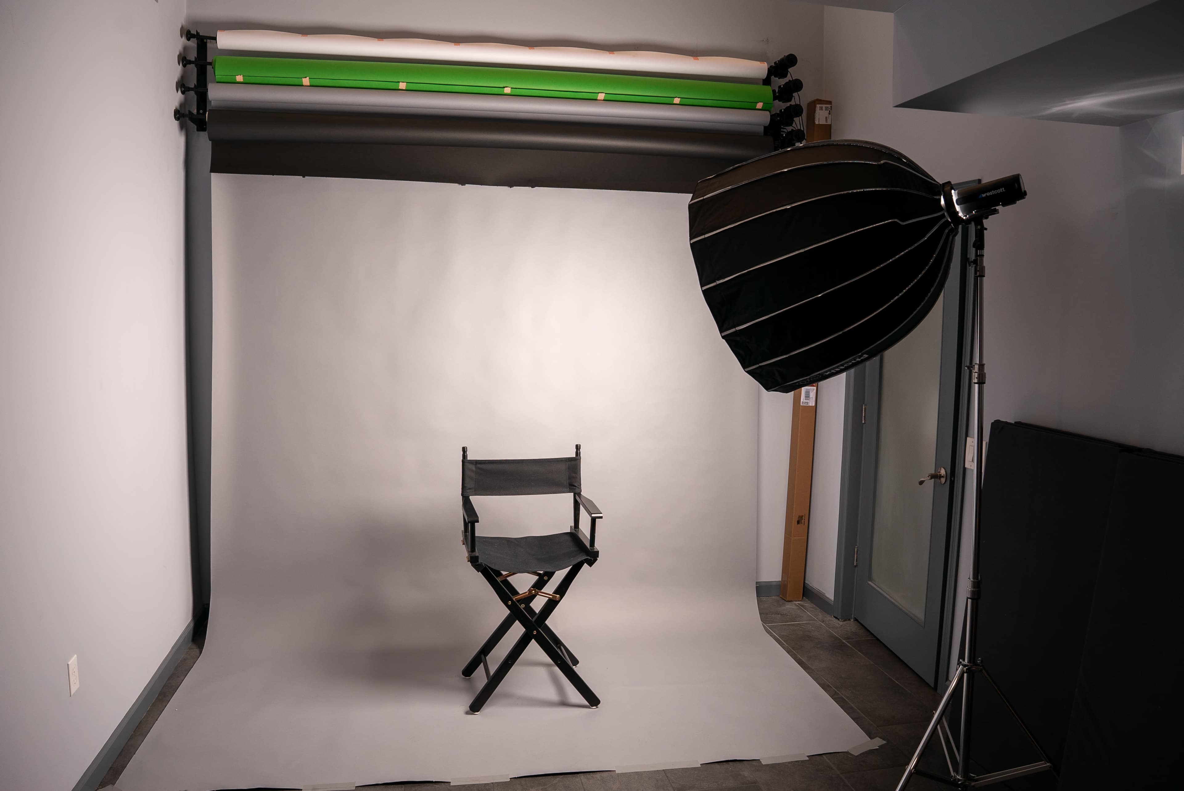 The image shows a photography studio with a folding chair positioned on a light backdrop, complemented by studio lighting equipment on the side.