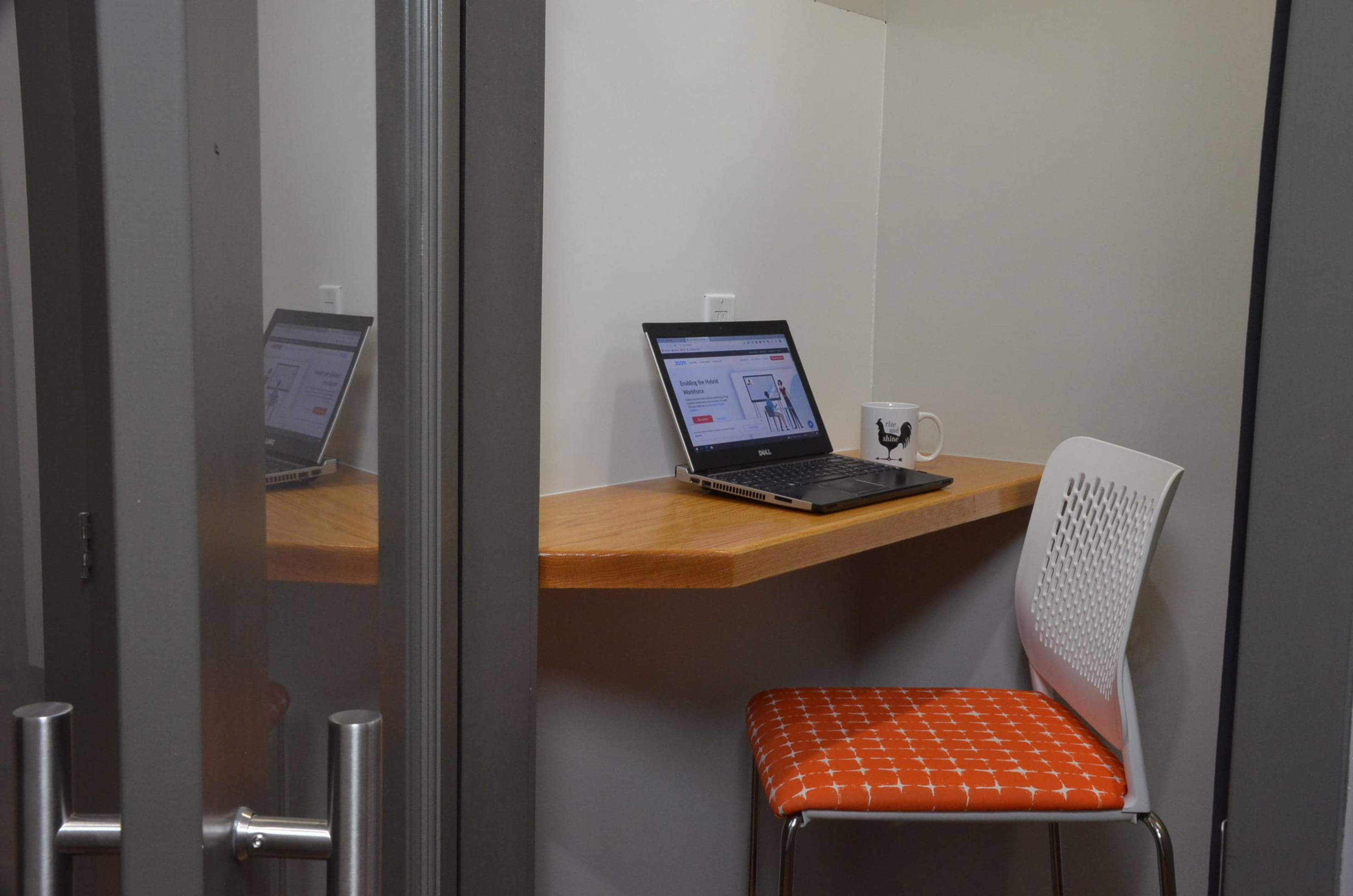 A small workspace features a laptop on a wooden shelf, a coffee mug, and a chair in a glass-walled room.