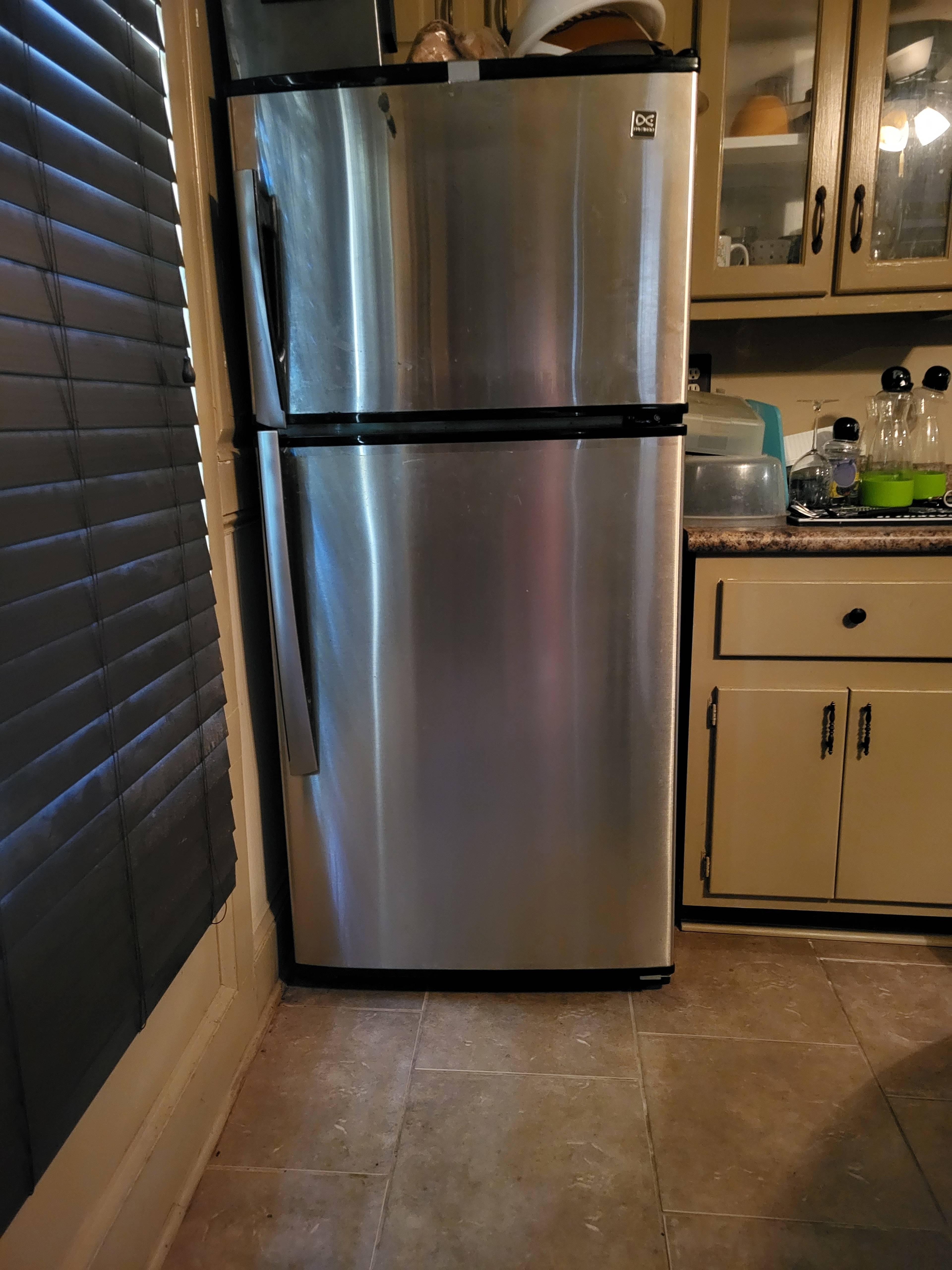 A metallic refrigerator stands against a wall in a kitchen with wooden cabinets and a tiled floor.