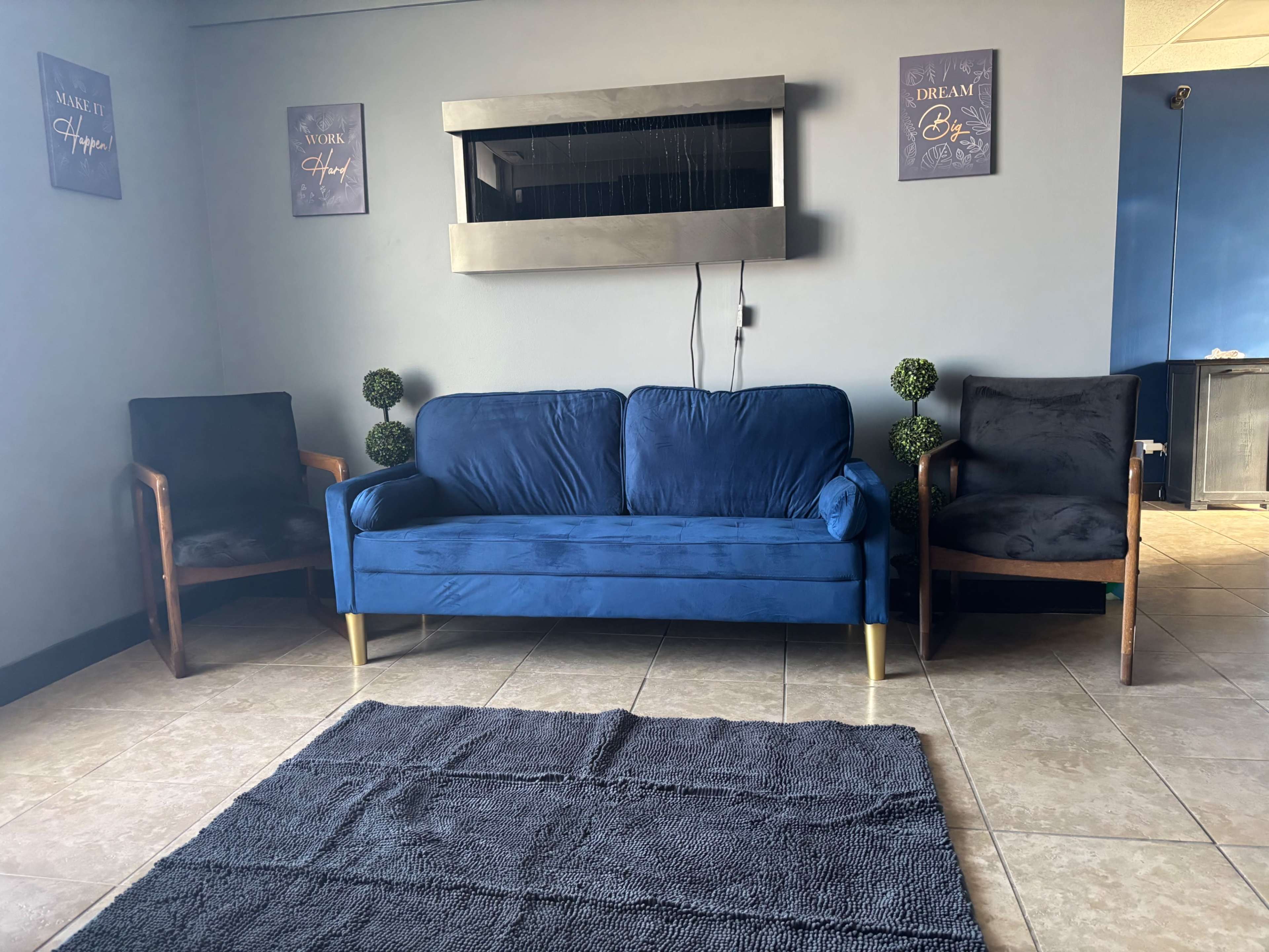 The image shows a seating area with a blue velvet couch, two wooden armchairs, and a textured rug on the tiled floor, against a wall adorned with framed motivational posters.