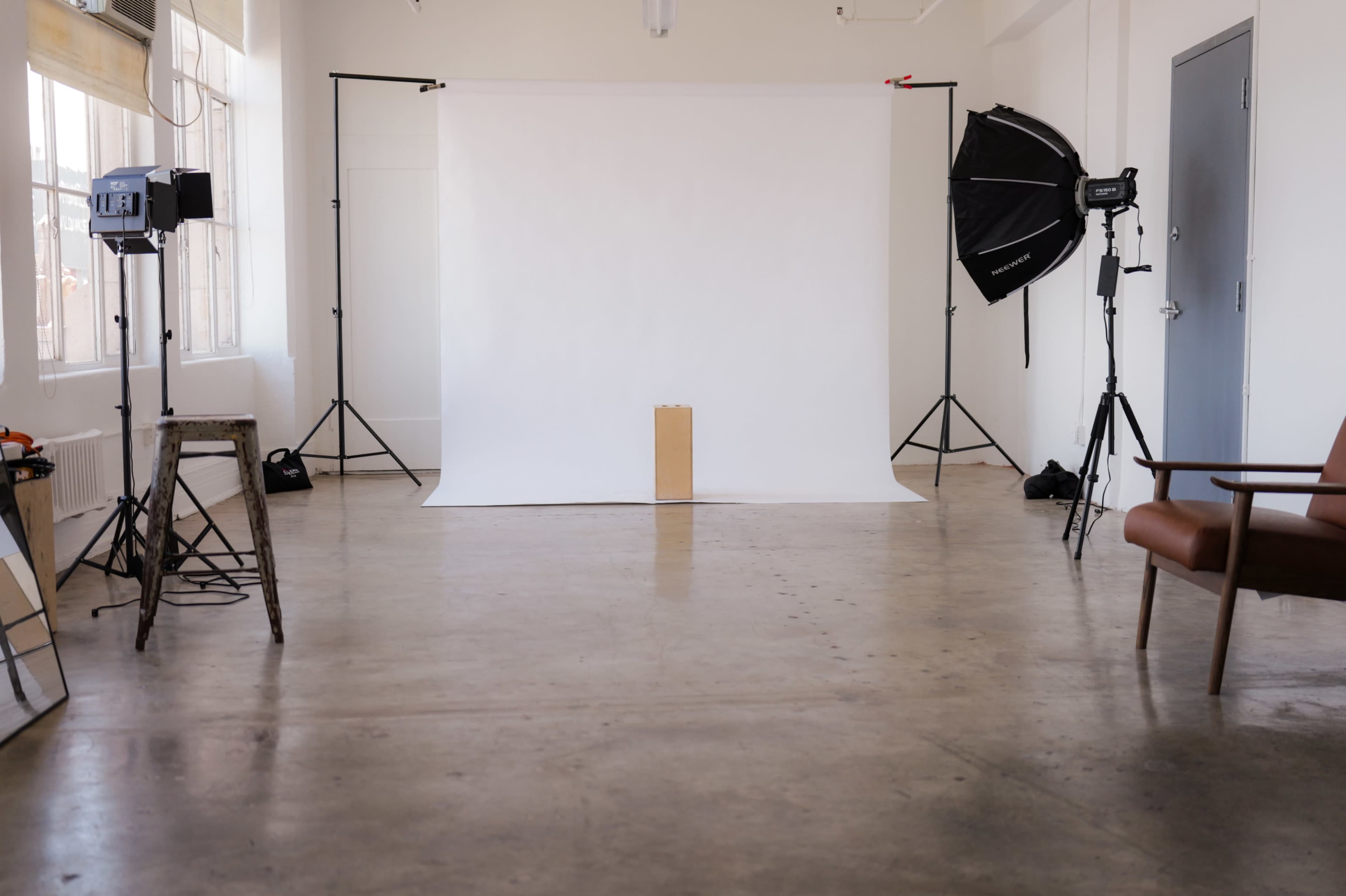 The image shows a photography studio with a plain white backdrop, a wooden block in front, and various lighting equipment arranged around the room.