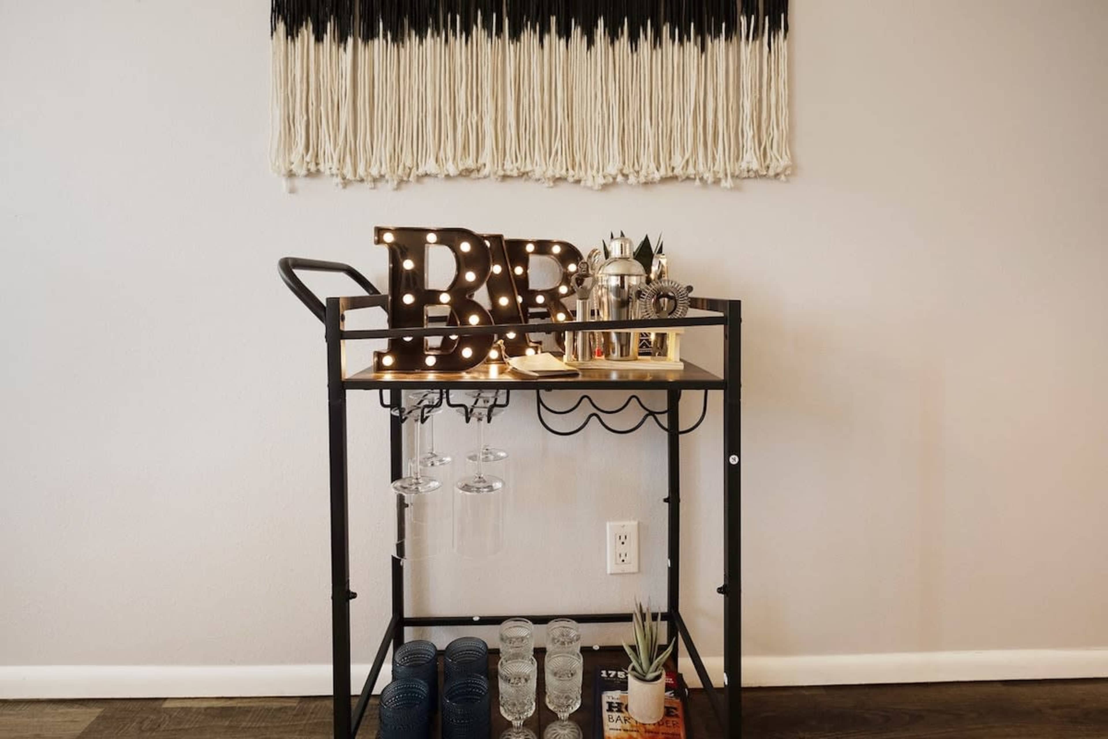 A bar cart features illuminated letters spelling "BAR" along with various glassware and decorative items beneath a wall hanging.