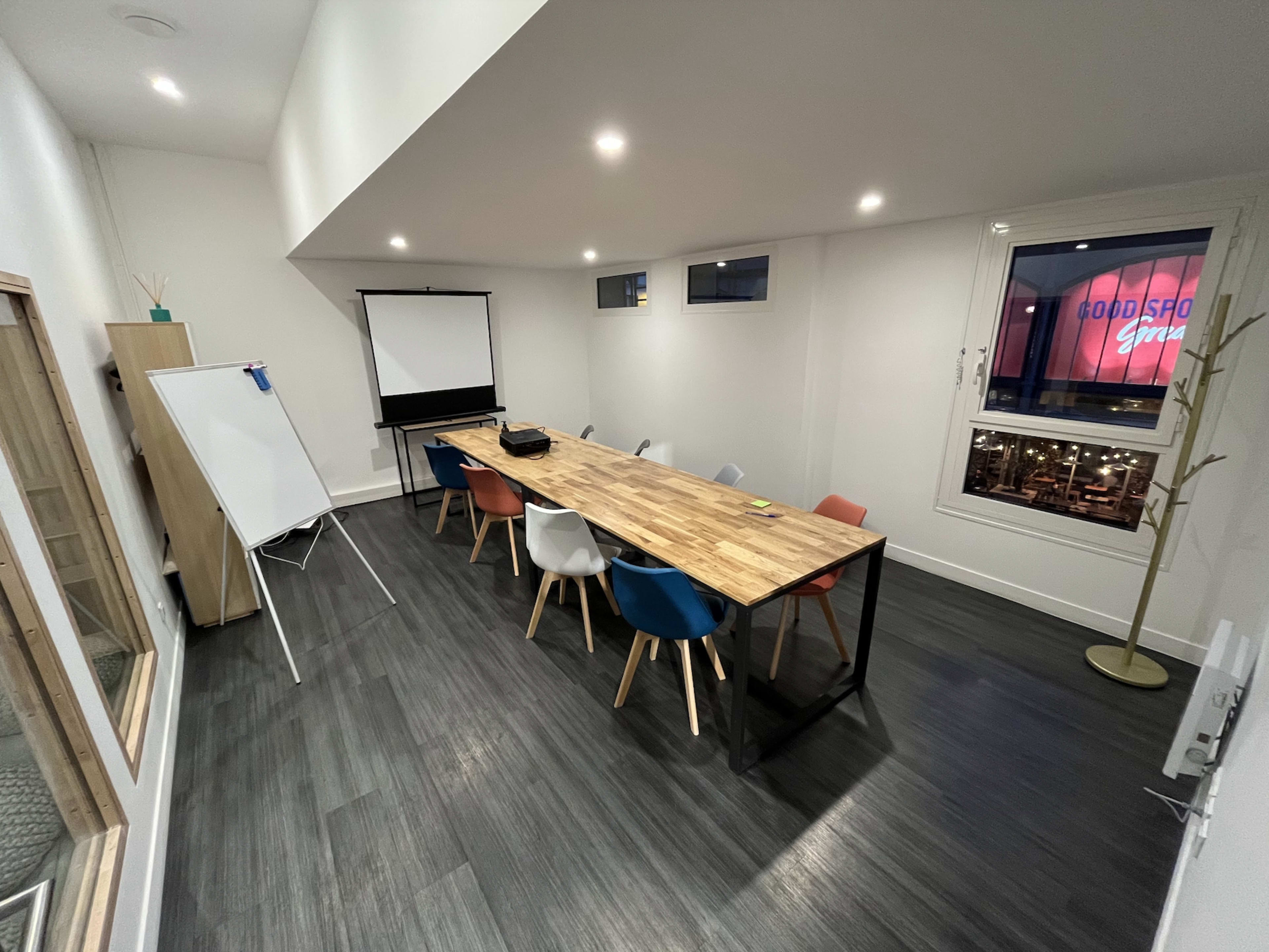 A wooden conference table is surrounded by colorful chairs in a bright, modern meeting room with a projector screen.