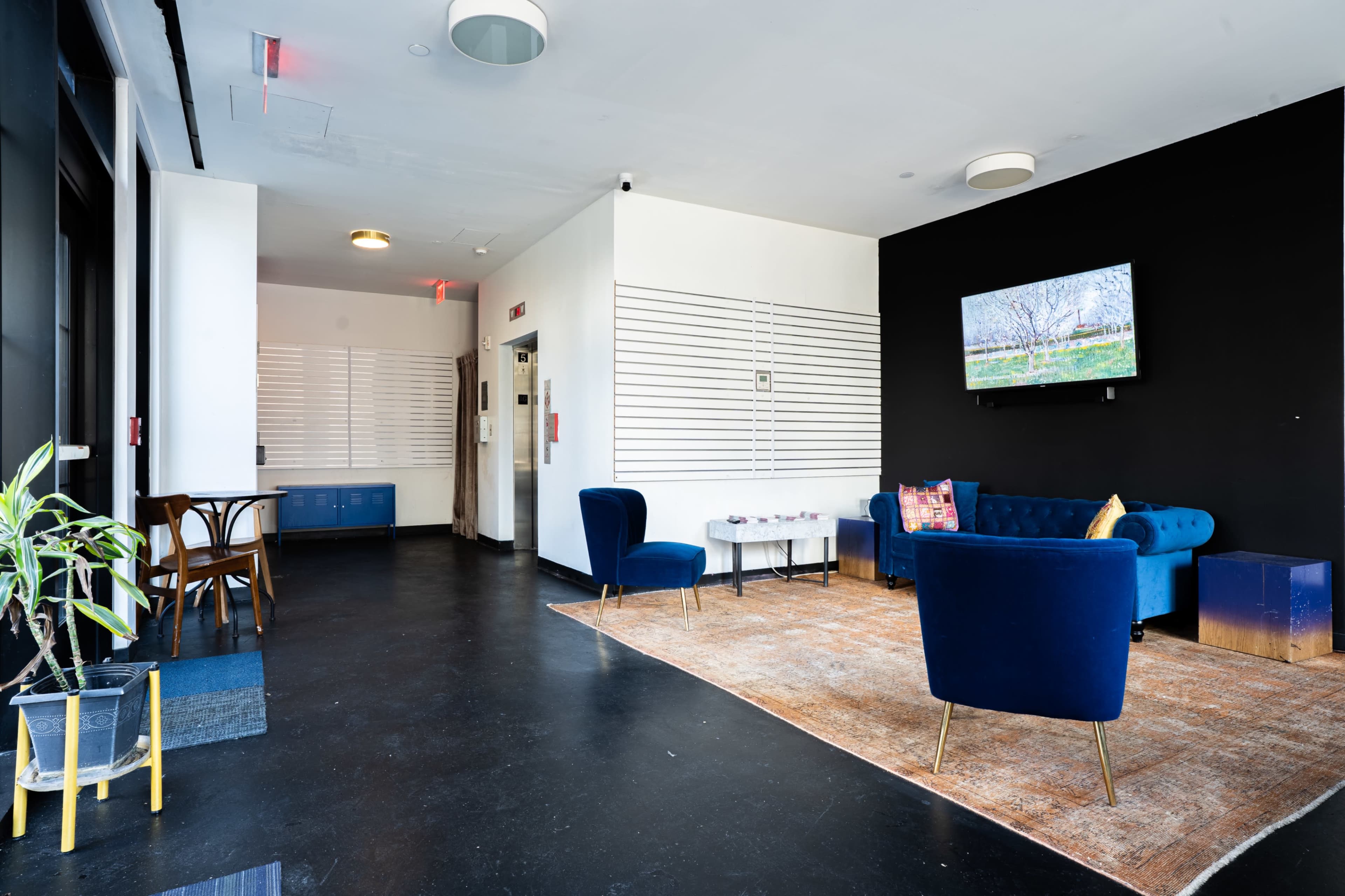 The image shows a modern lounge area with a blue sofa, two blue chairs, a coffee table, and a television mounted on the black wall.