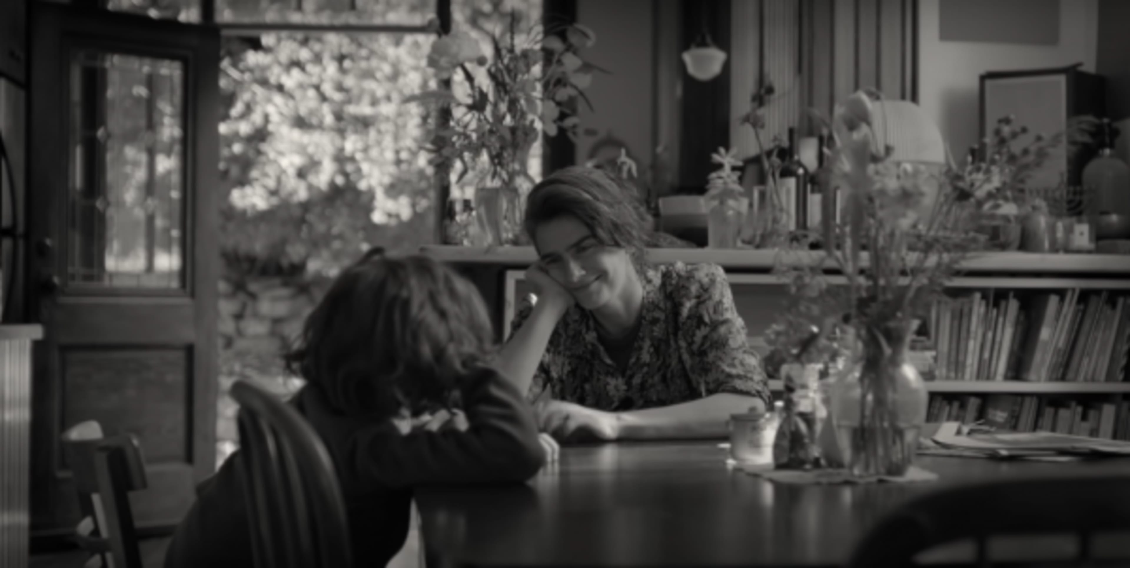 A woman smiles at a child seated at a wooden table in a sunlit room filled with plants and books.