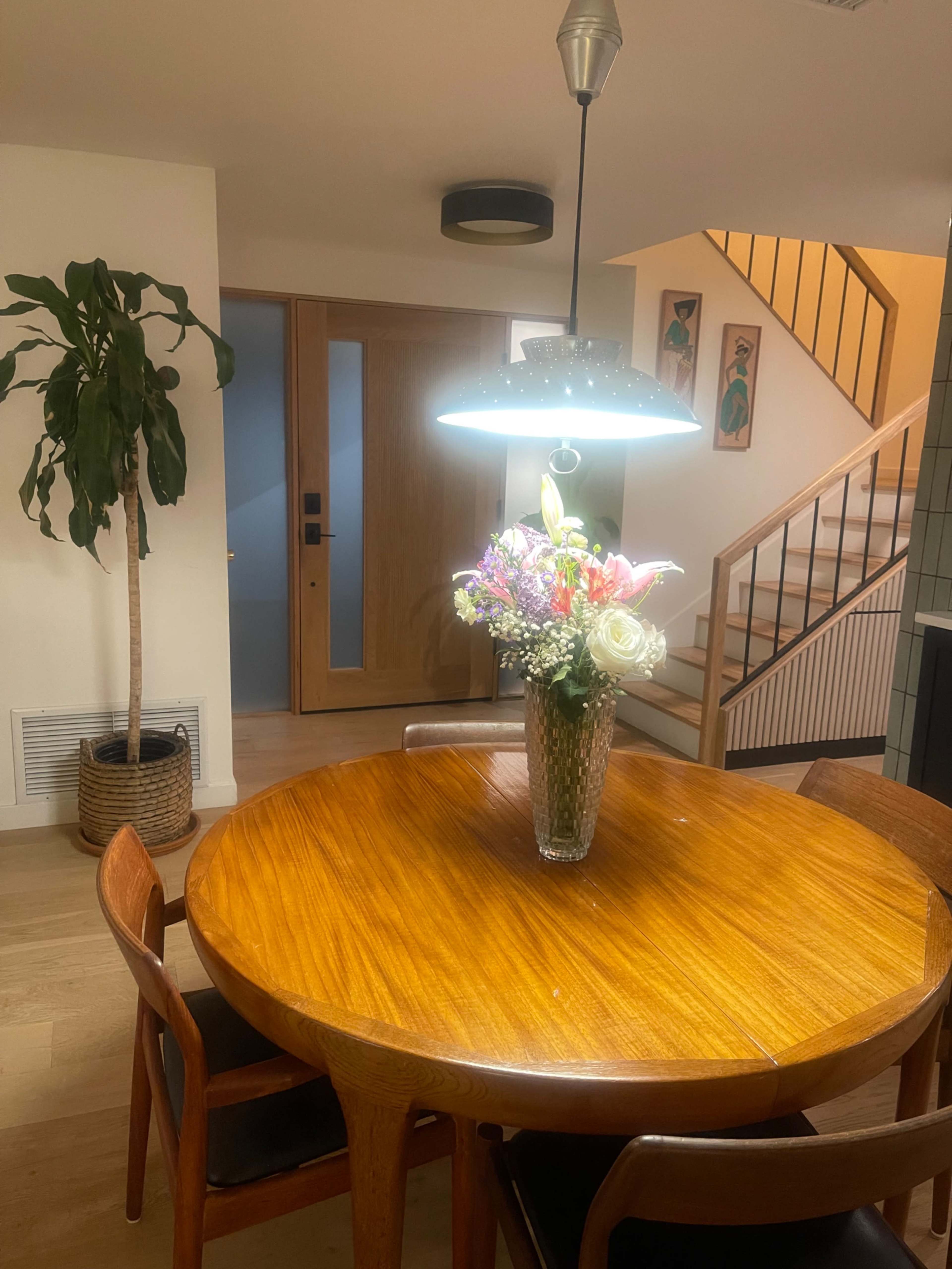 A wooden dining table with chairs is centered under a pendant light, adorned with a vase of colorful flowers, while a plant stands to the side and a staircase is visible in the background.