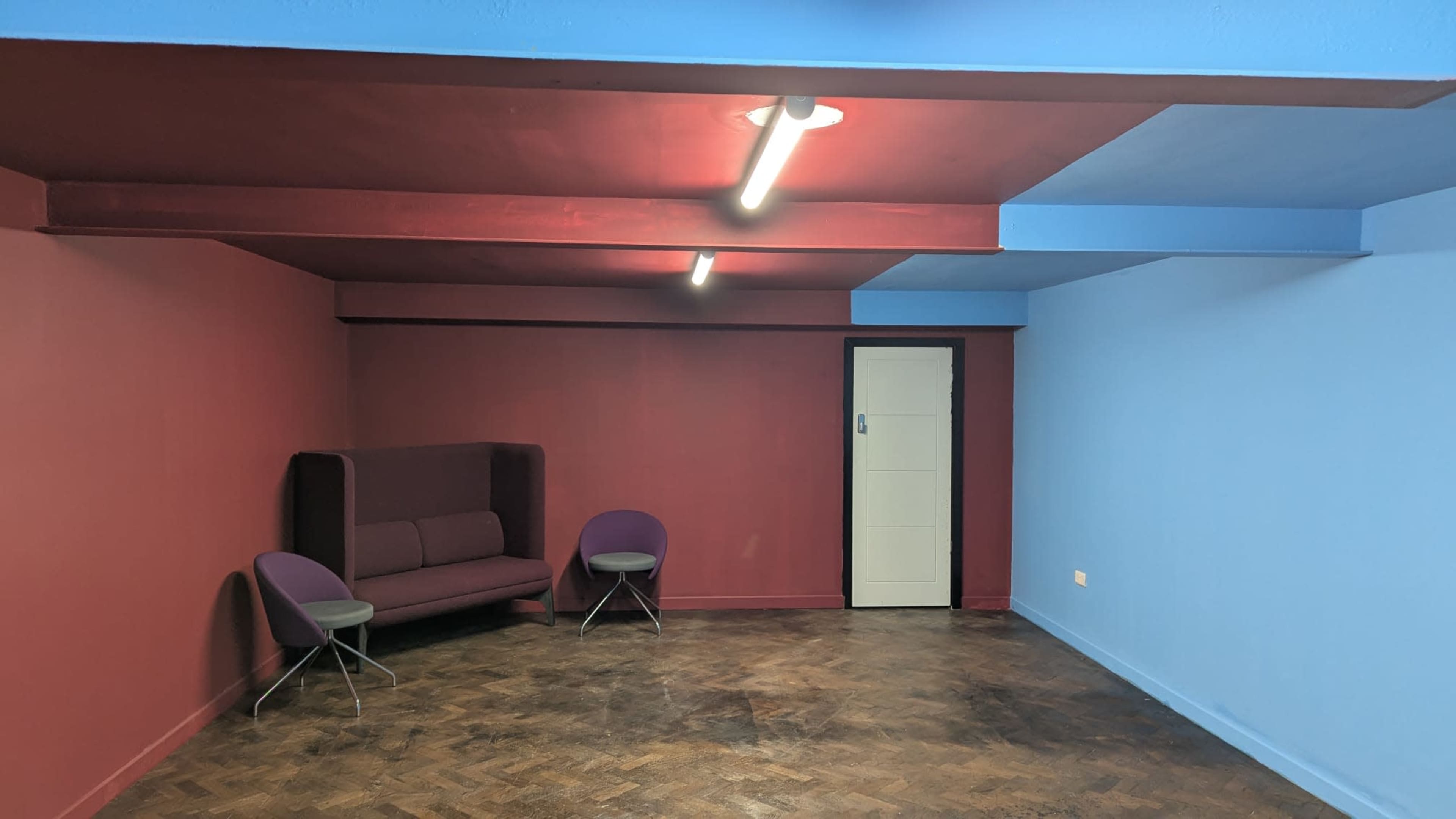 The image shows an empty room with maroon and blue walls, a dark purple couch, and two gray chairs near a white door.