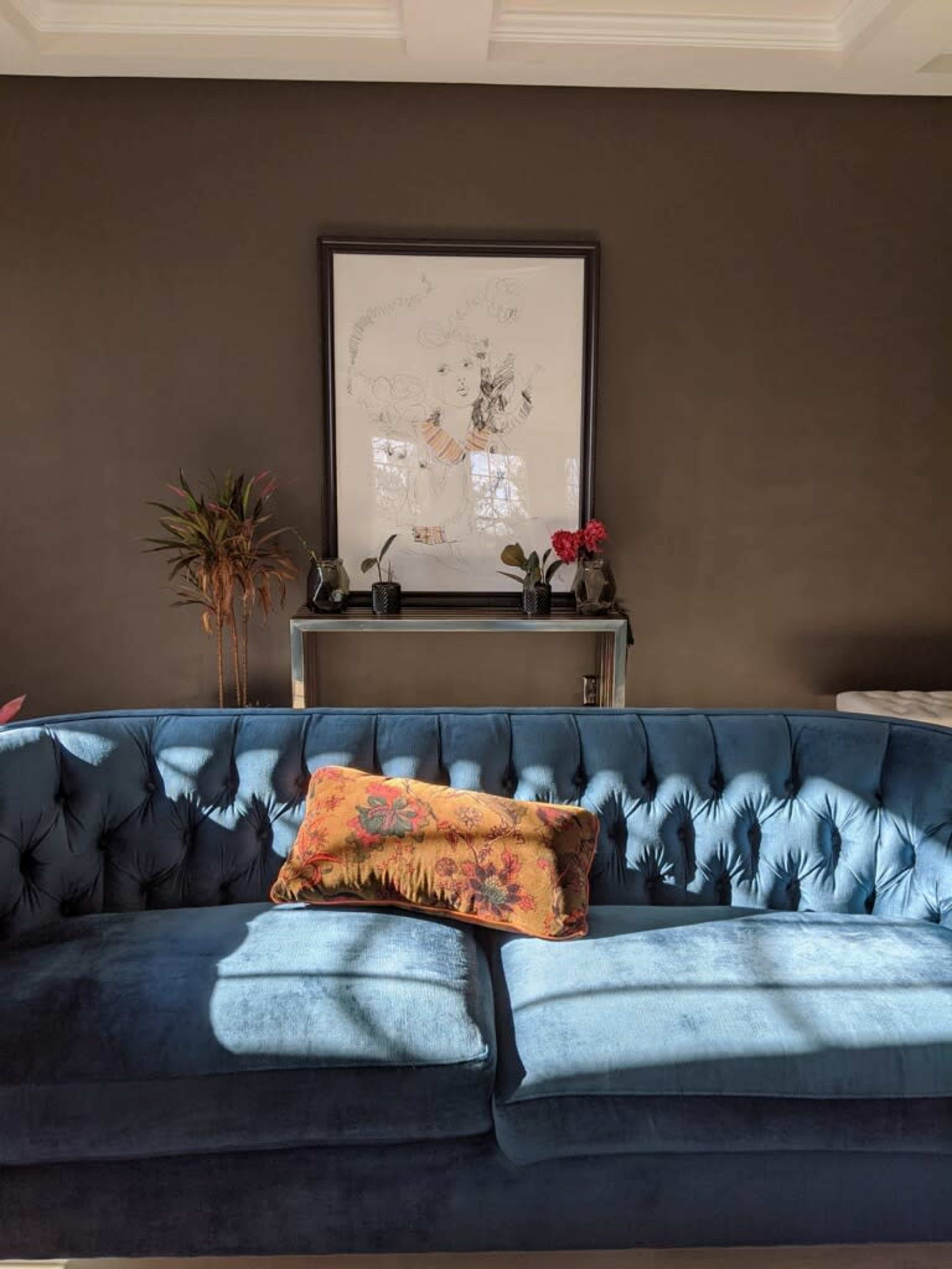 A blue tufted sofa in a room with dark walls, a framed artwork above it, and a small table with potted plants and flowers.