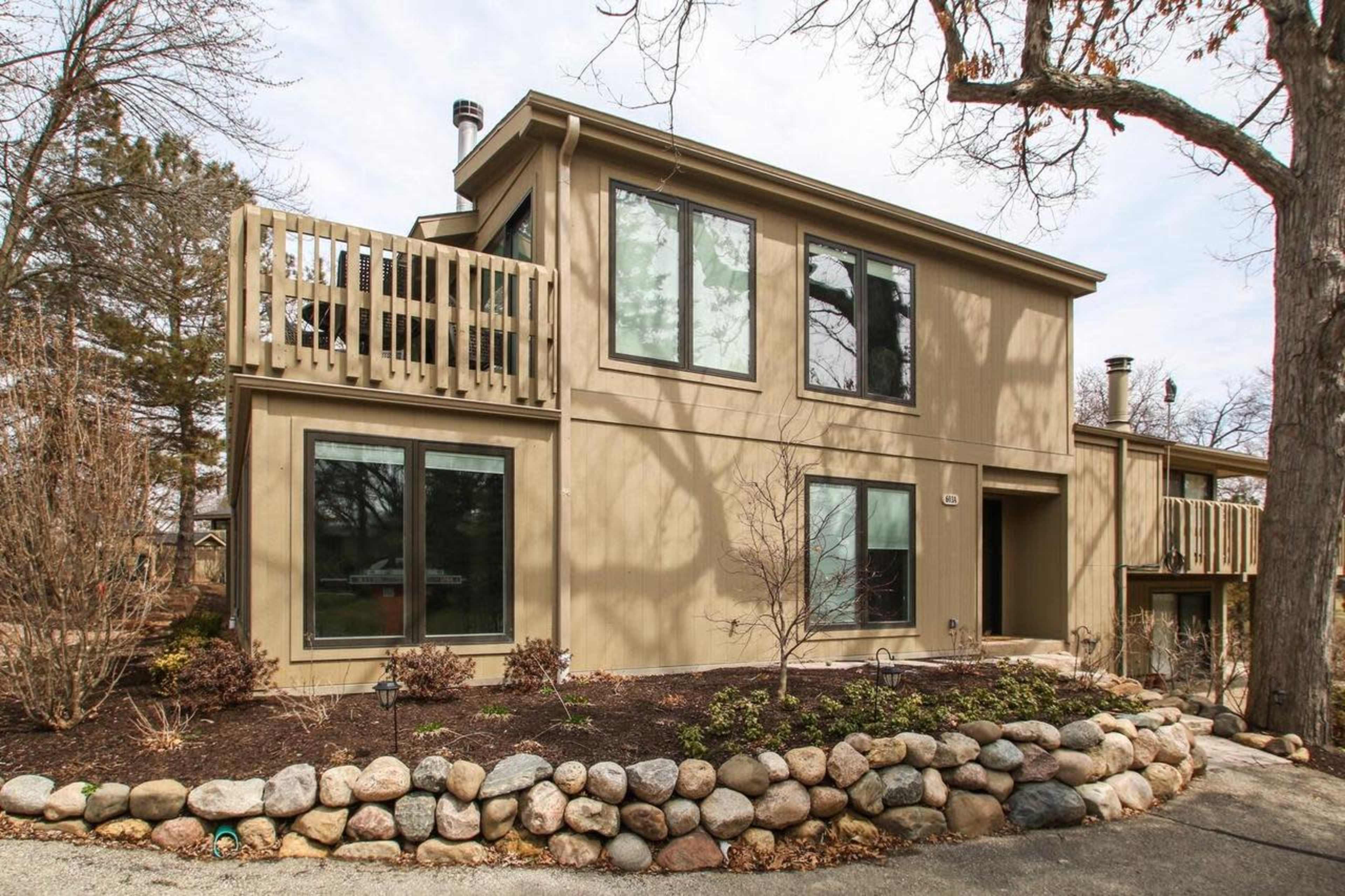 A two-story house with large windows, a balcony, and a landscaped yard featuring a stone border and bare trees.