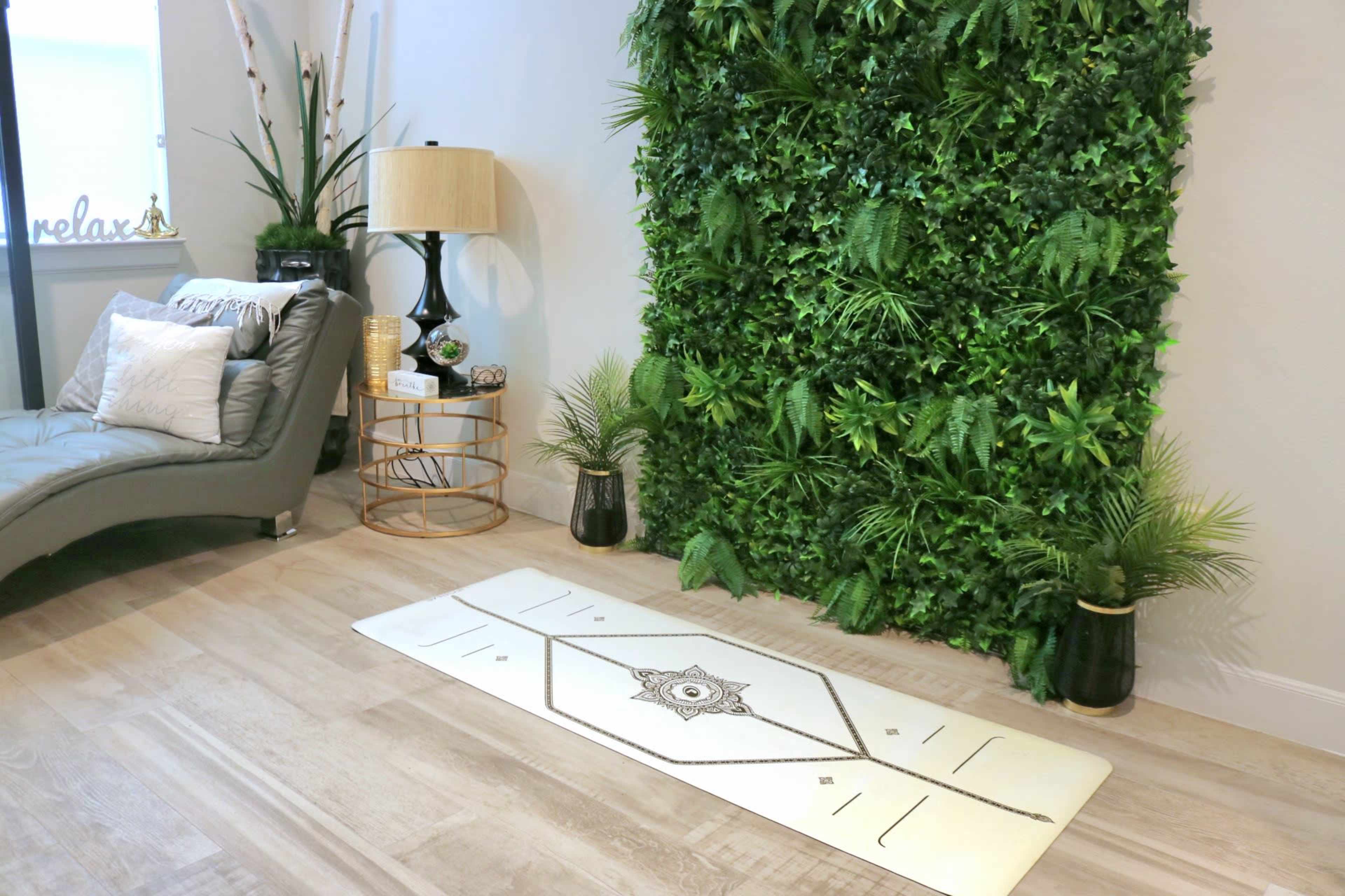 The scene features a modern living space with a green wall of plants, a decorative rug with a geometric pattern, and a gray chaise lounge beside a lamp and small table.