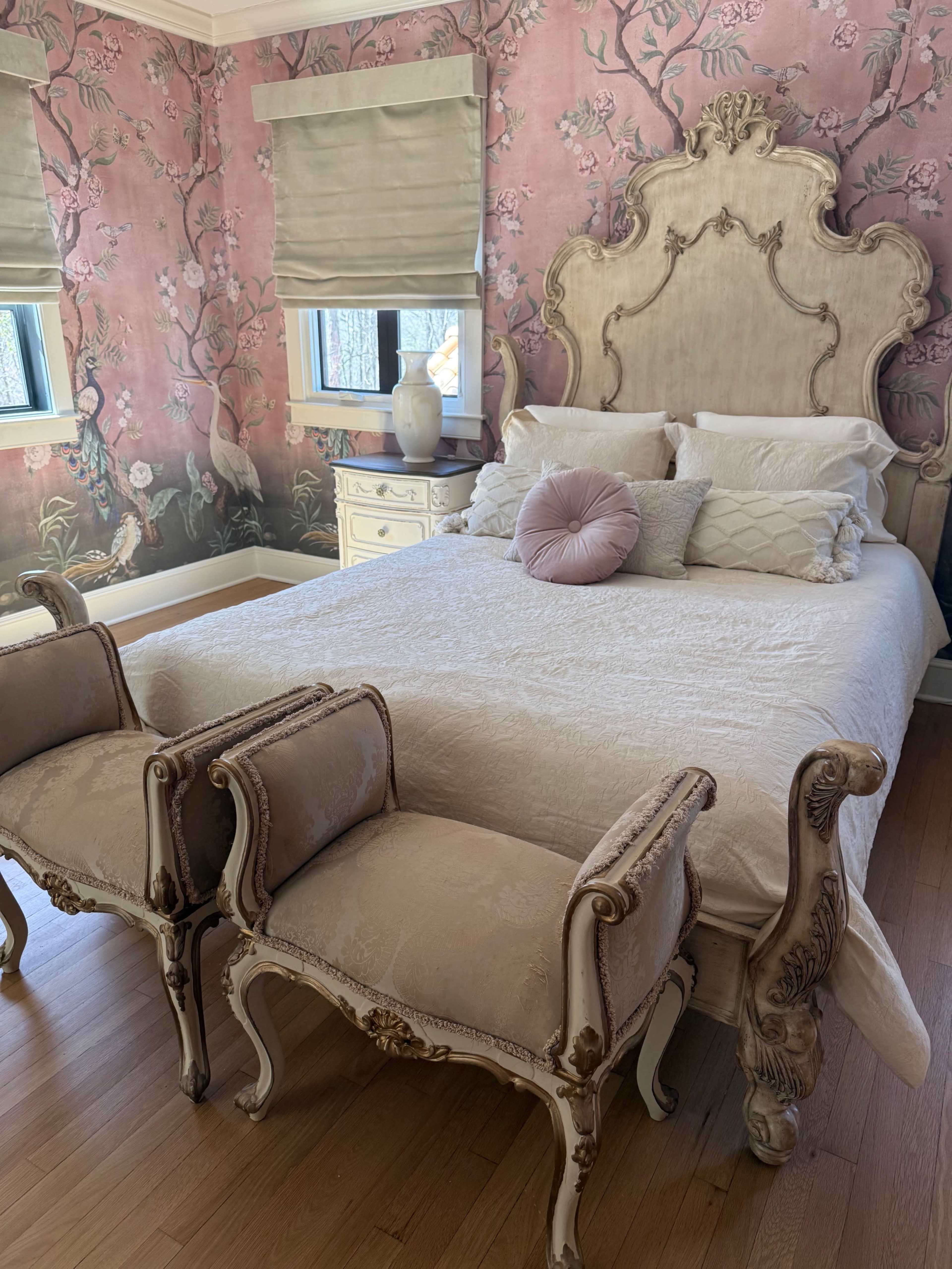 The image shows a beautifully decorated bedroom featuring a large, ornate bed with a light-colored comforter and two decorative chairs at the foot of the bed, set against floral wallpaper.