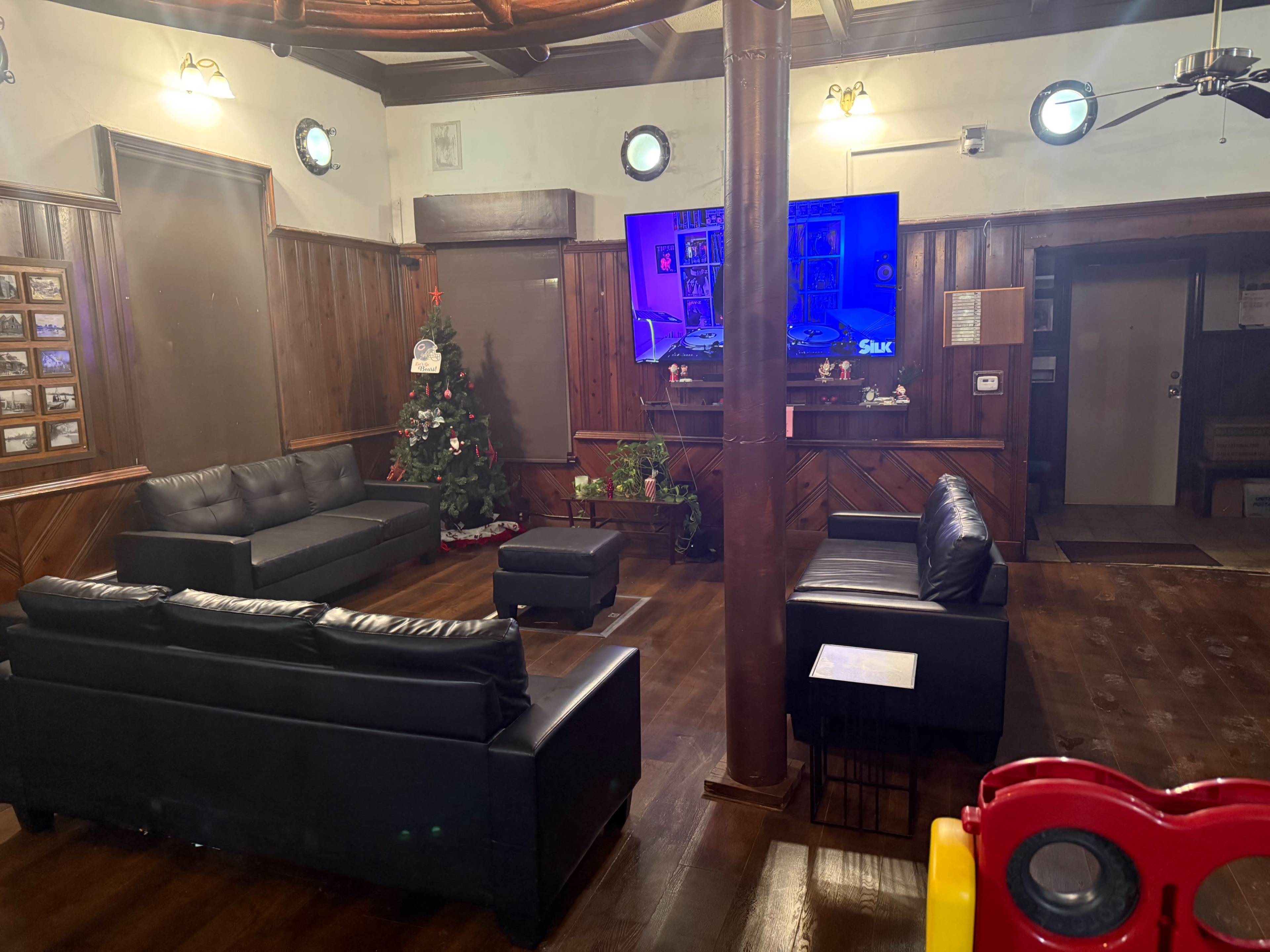 The image shows a cozy lounge area with black leather furniture, a Christmas tree, and a television mounted on a wooden wall.