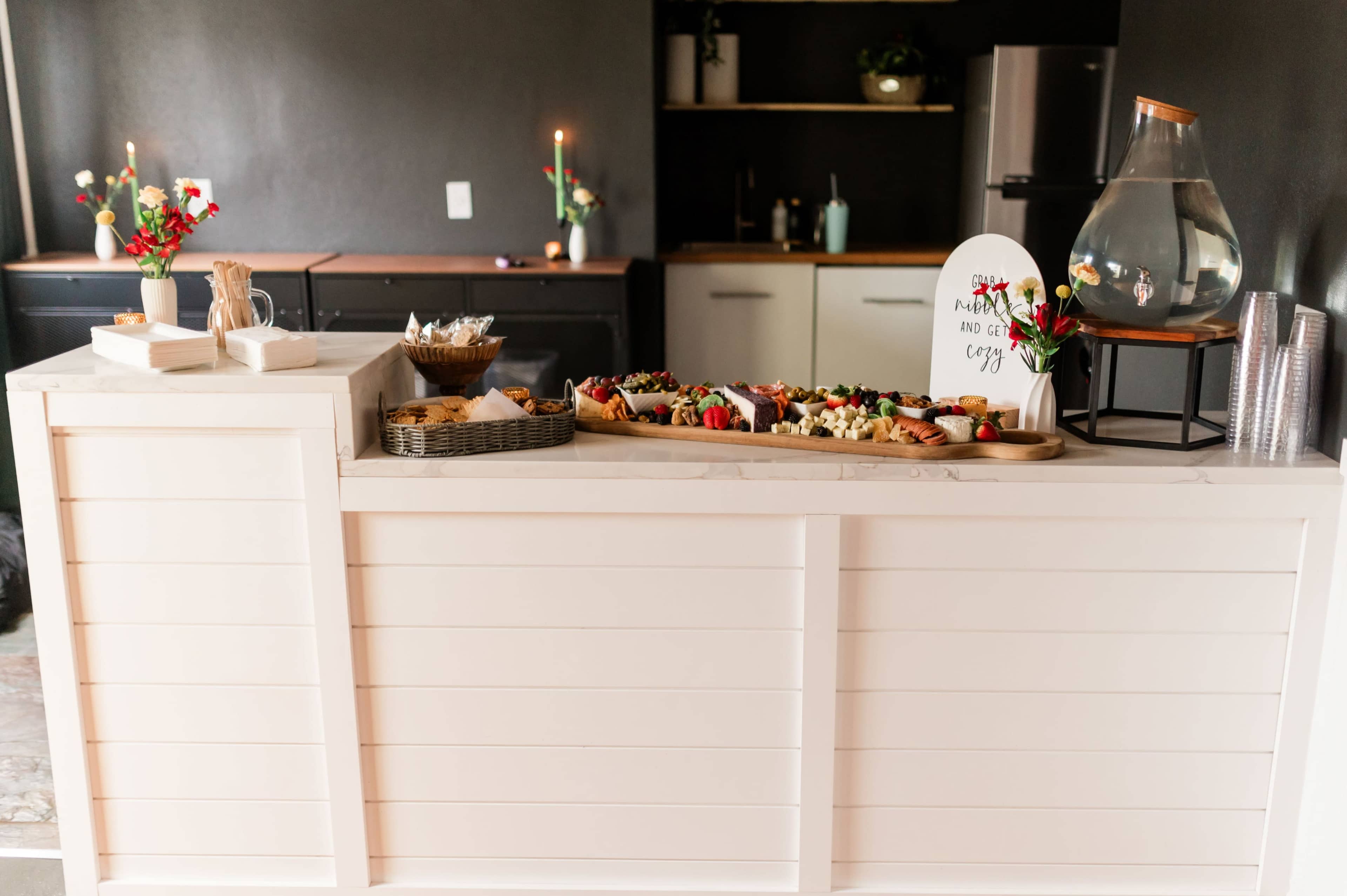 A refreshment station displays a variety of food items on a countertop, with a decorative sign and beverages in a clear container.