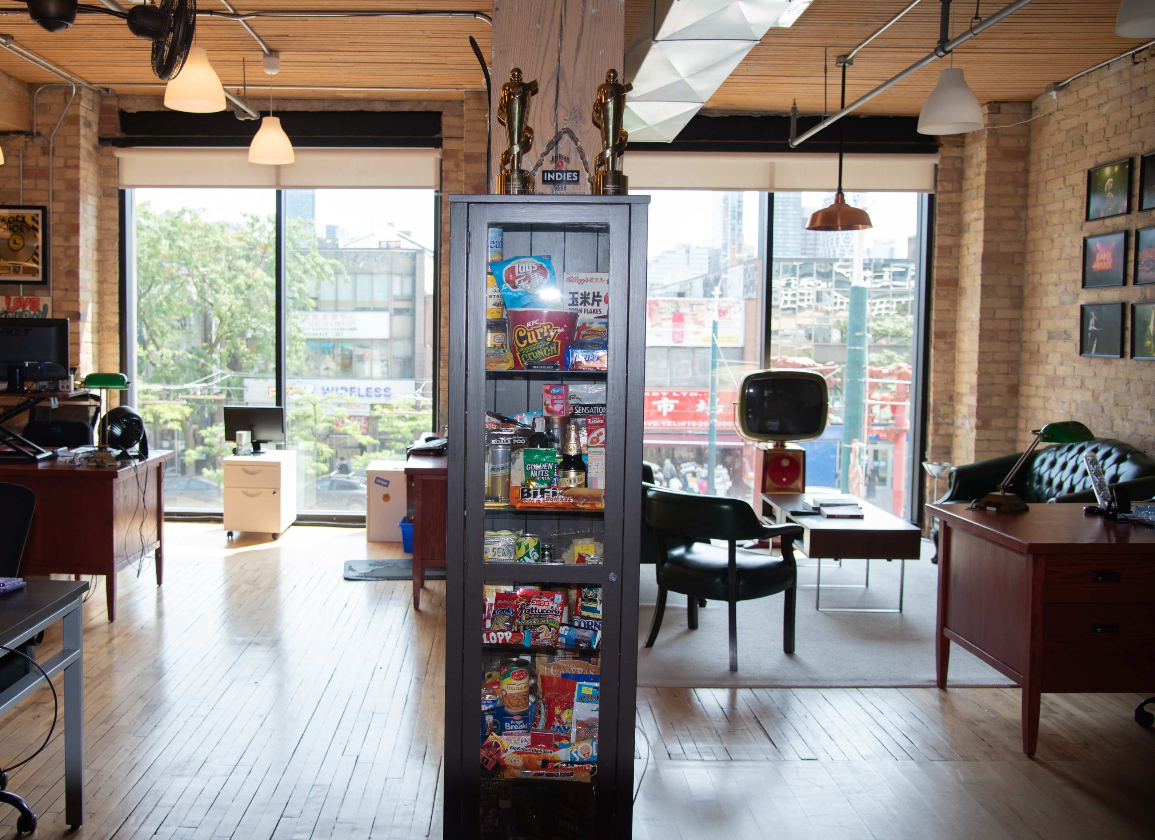The image shows an office interior featuring a display cabinet filled with snacks, wooden desks, and large windows overlooking an urban landscape.