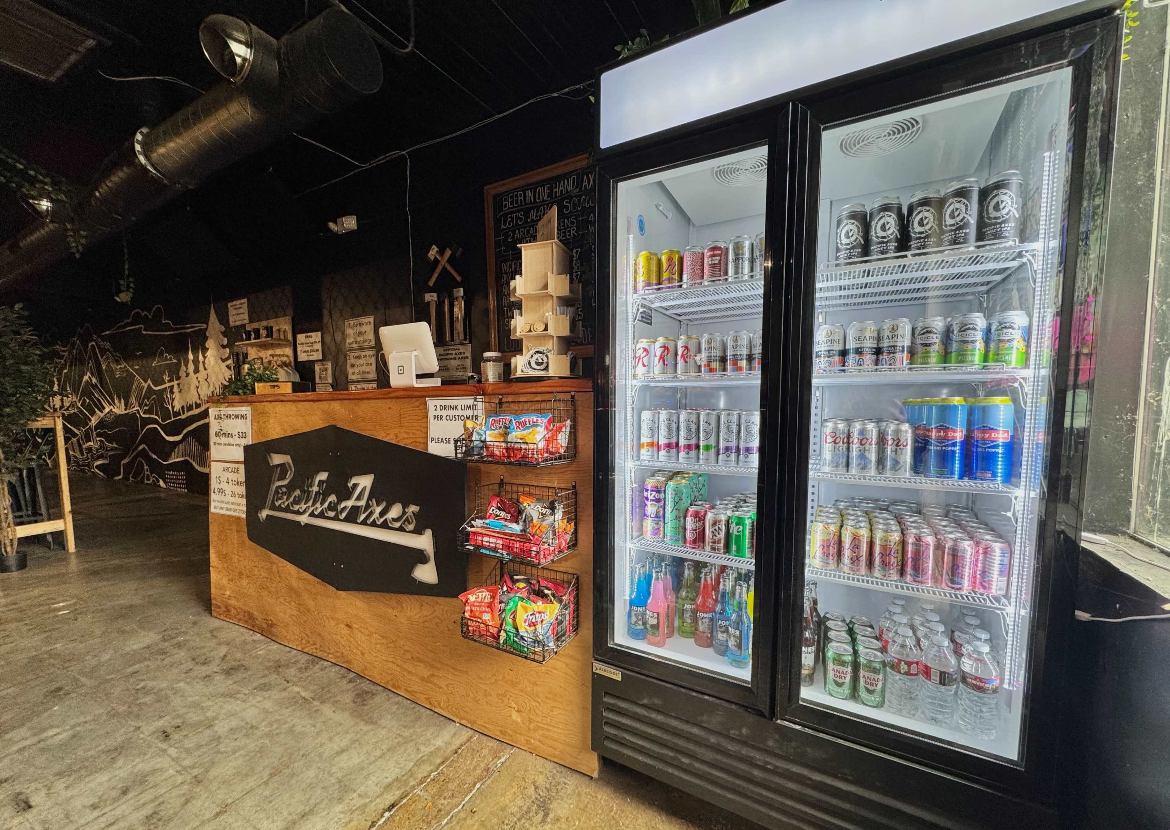 The image shows a corner of a shop with a wooden counter displaying various snack items and a refrigerator filled with colorful drinks.