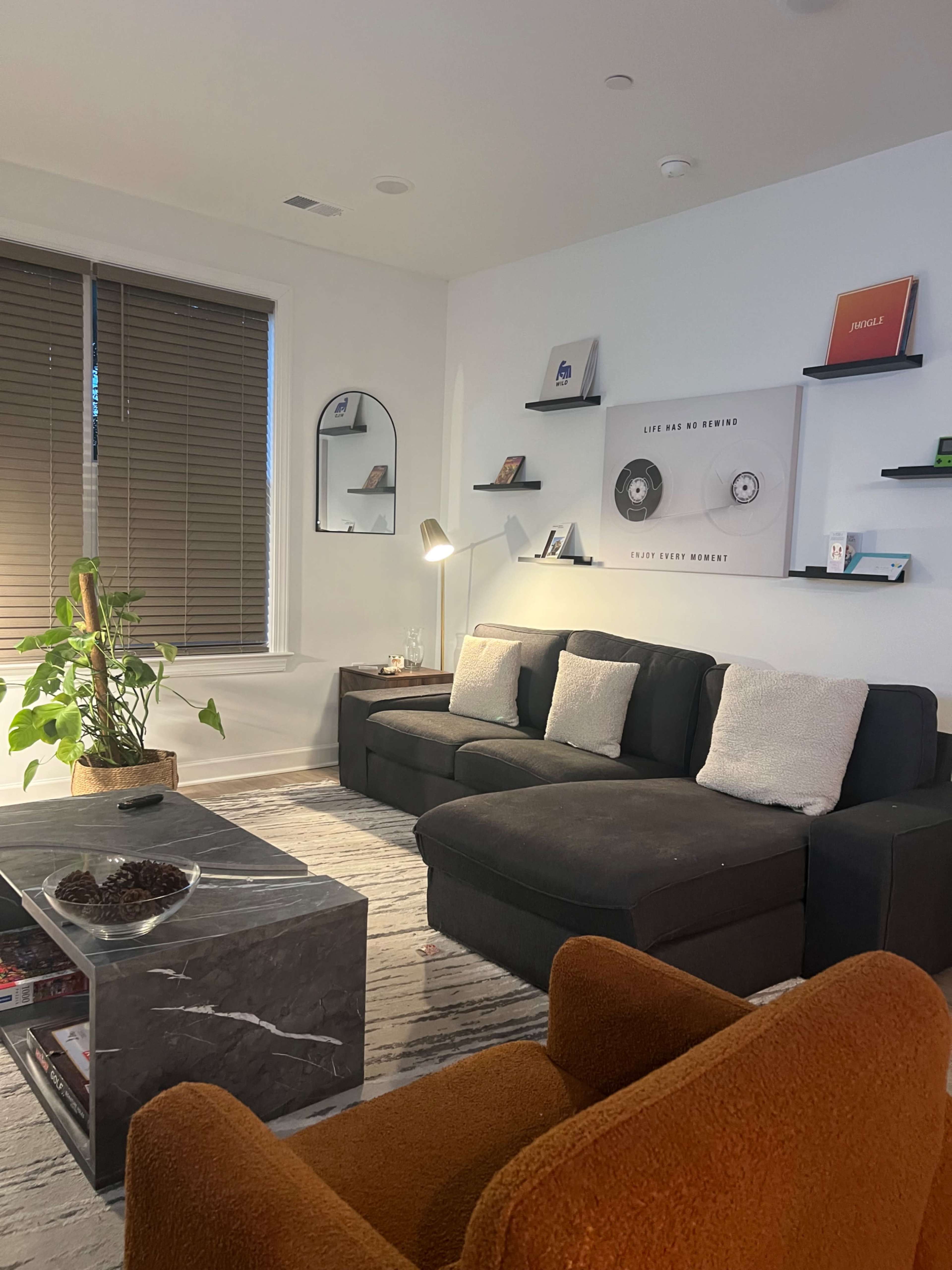 The living room features a black sectional sofa, an orange armchair, a marble coffee table, and a large window with blinds.