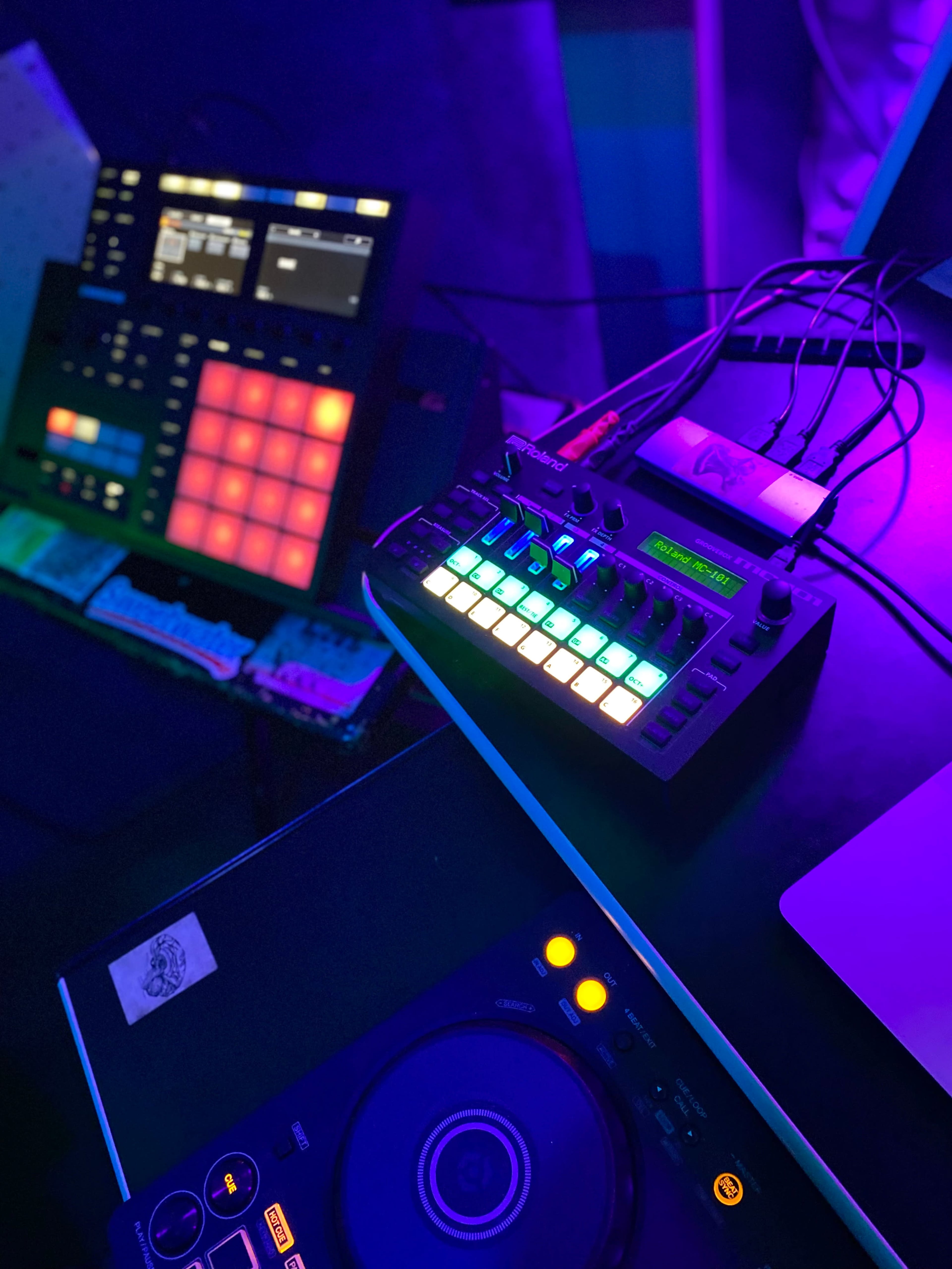 The image displays a close-up view of a DJ setup with colorful illuminated pads and a mixer, surrounded by various electronic devices and cables.