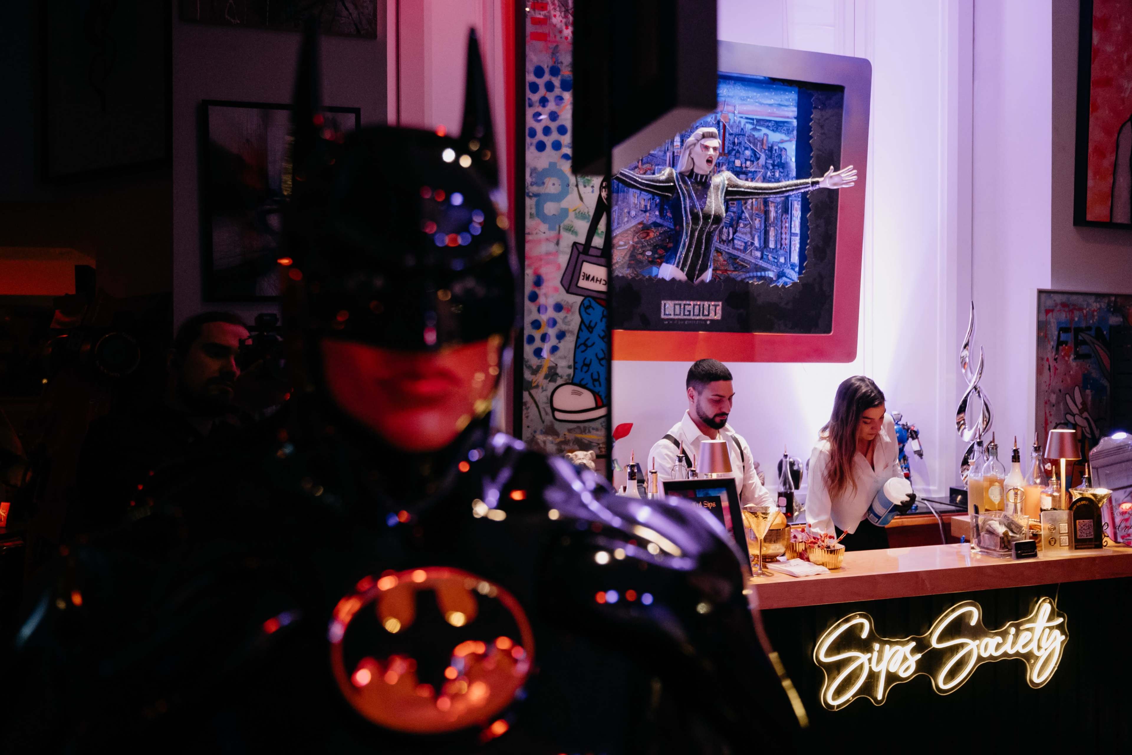 A Batman statue stands prominently in the foreground, while a bar with illuminated signage and patrons is visible in the background.