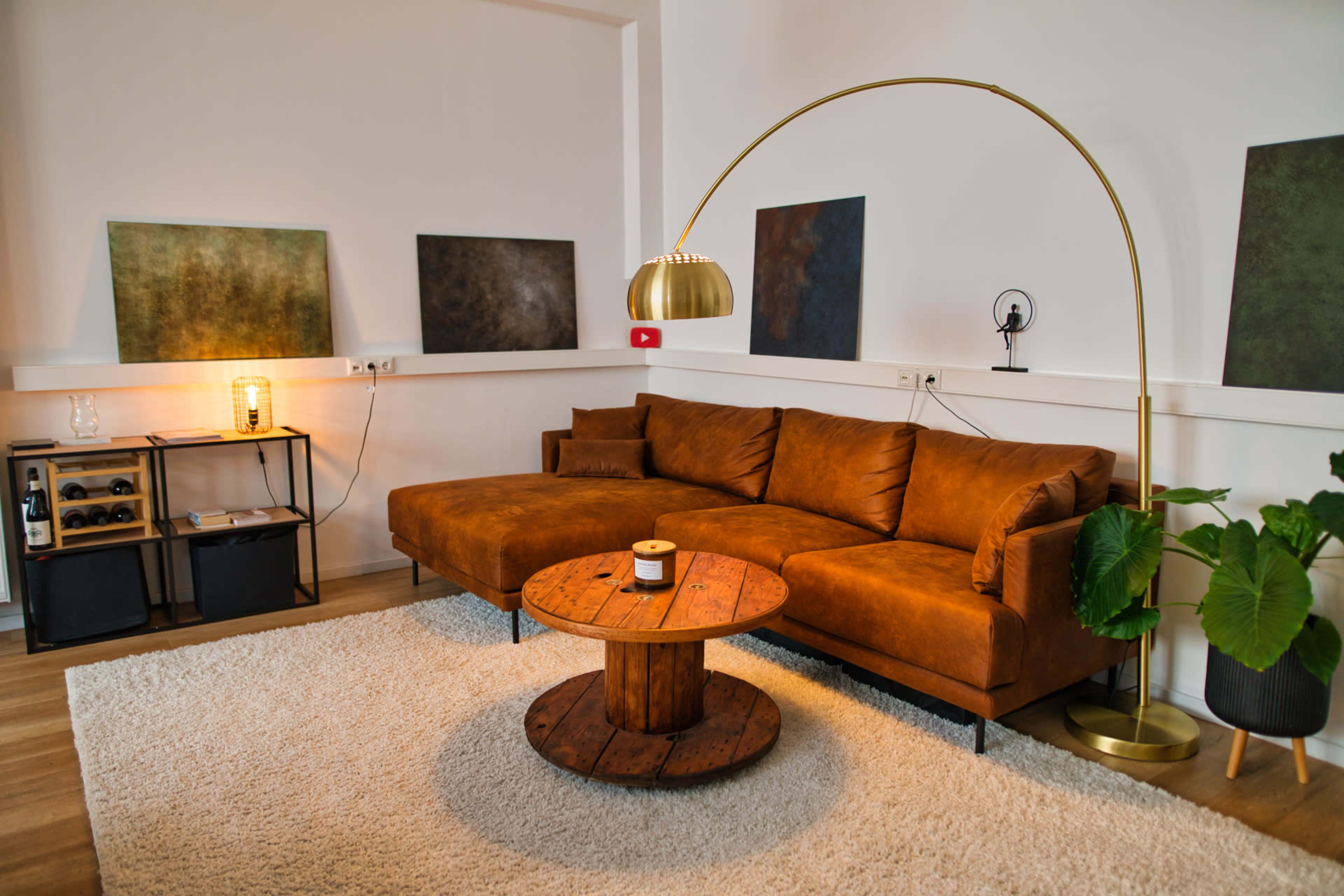 The image shows a modern living room featuring a brown sectional sofa, a round wooden coffee table, a floor lamp, and abstract wall art.