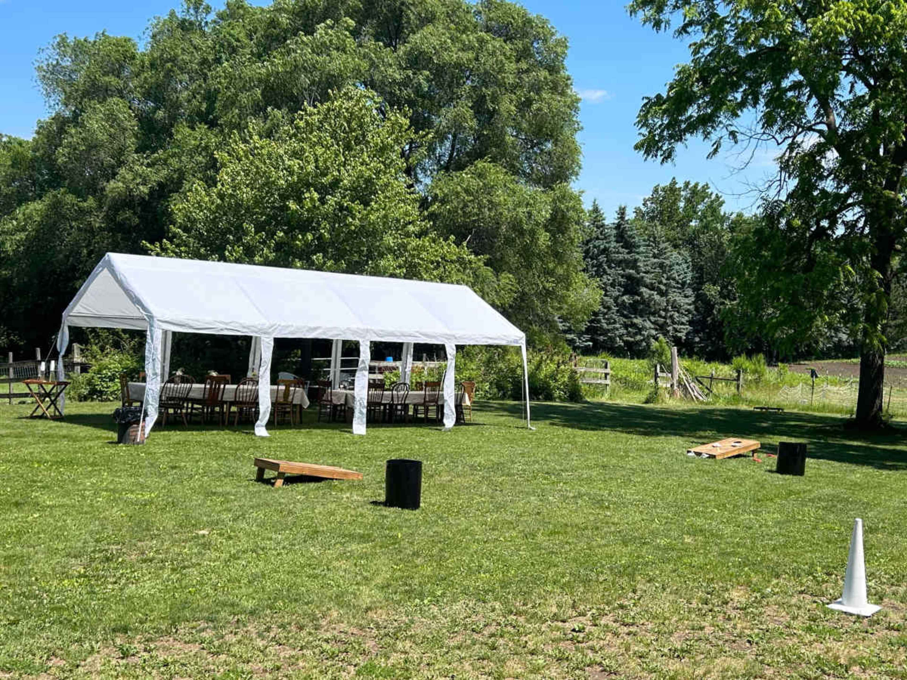 A large white tent is set up on a grassy area, surrounded by trees and featuring tables and chairs inside, with cornhole boards placed nearby.