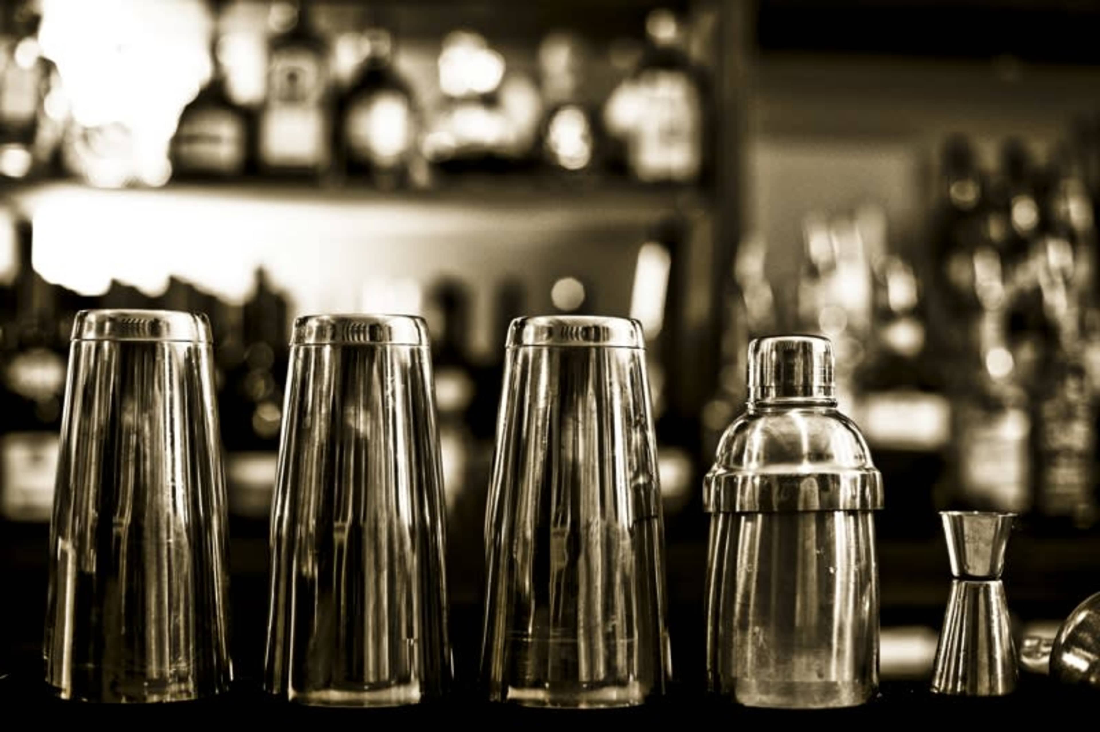 A row of silver cocktail shakers and measuring jigger set against a blurred bar background filled with bottles.