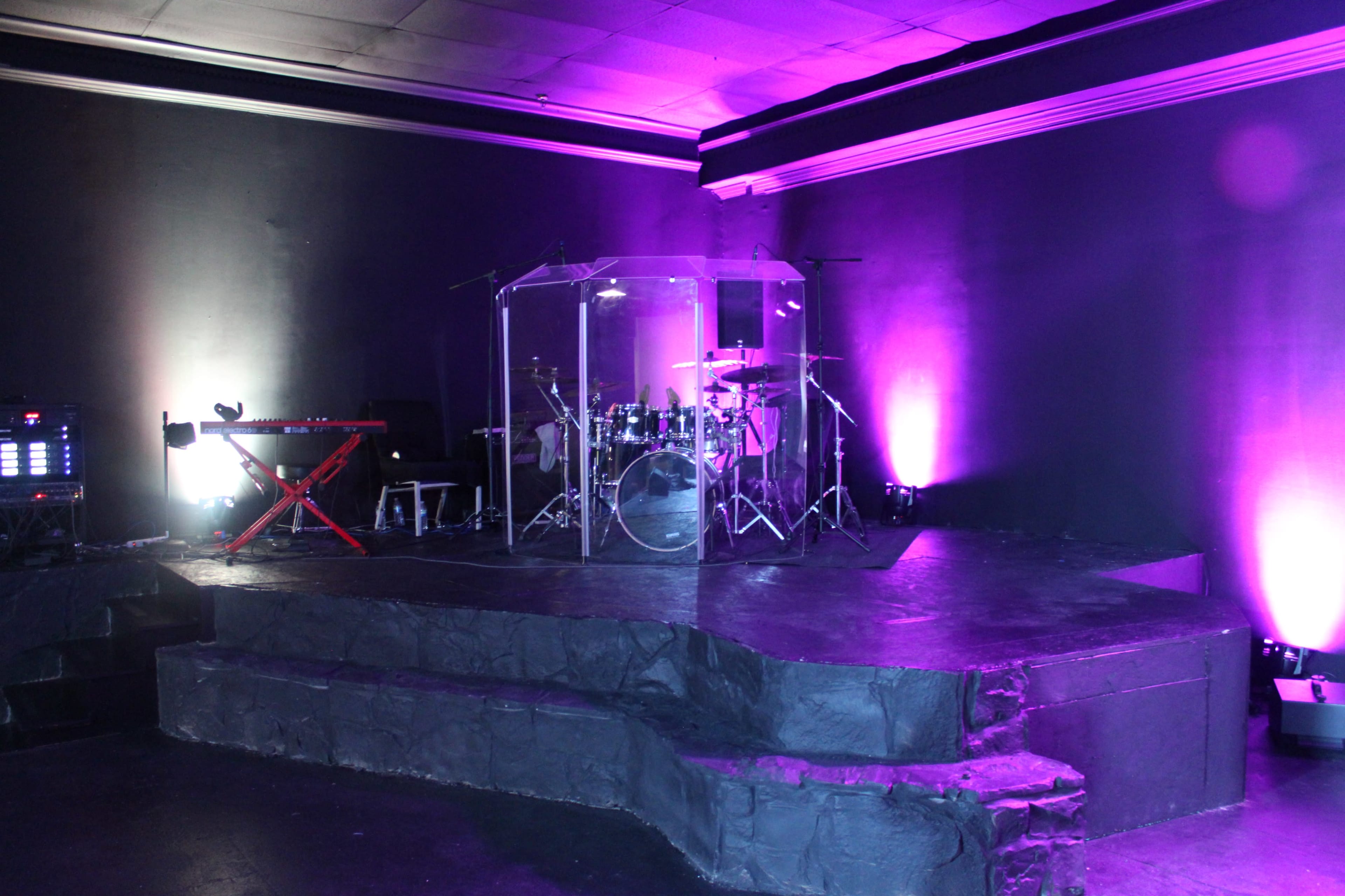 The image shows a stage with a drum set enclosed in a transparent sound shell, flanked by a keyboard and illuminated with purple lighting.