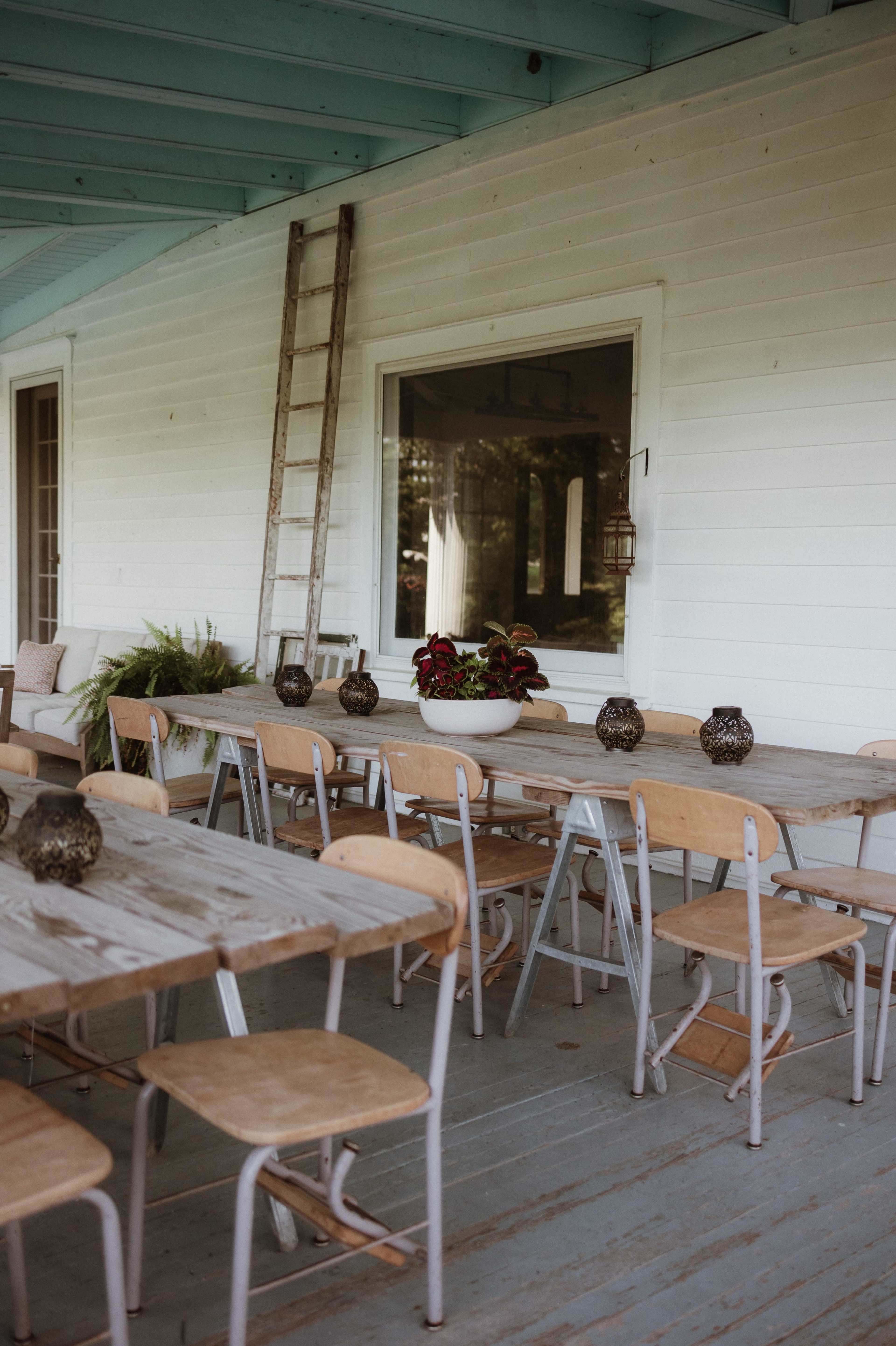 A porch with several wooden tables and chairs arranged neatly, along with a ladder against the wall and decorative items on the tables.