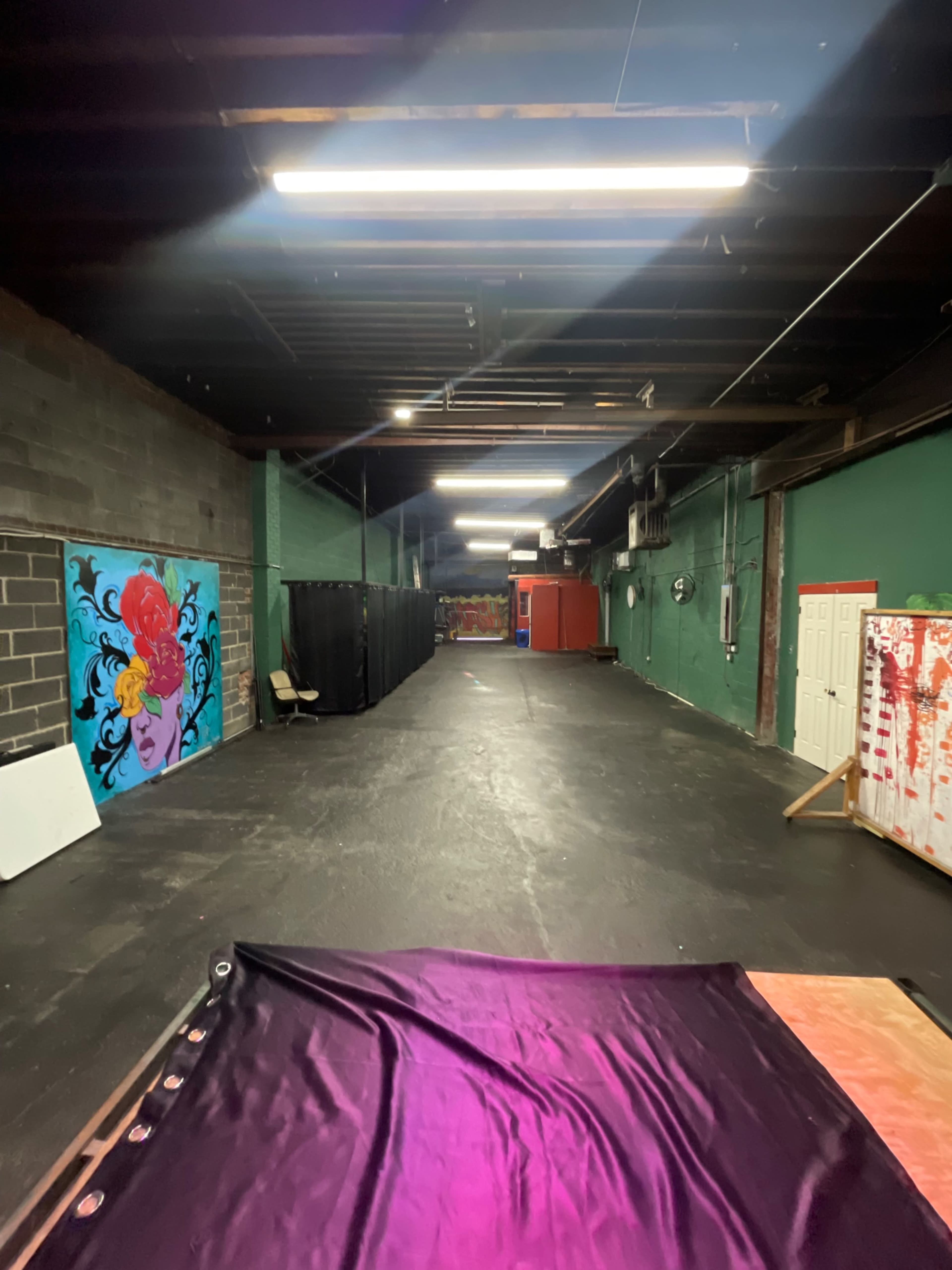 The image shows a long, narrow warehouse space with colorful murals on the walls and various items along the floor.