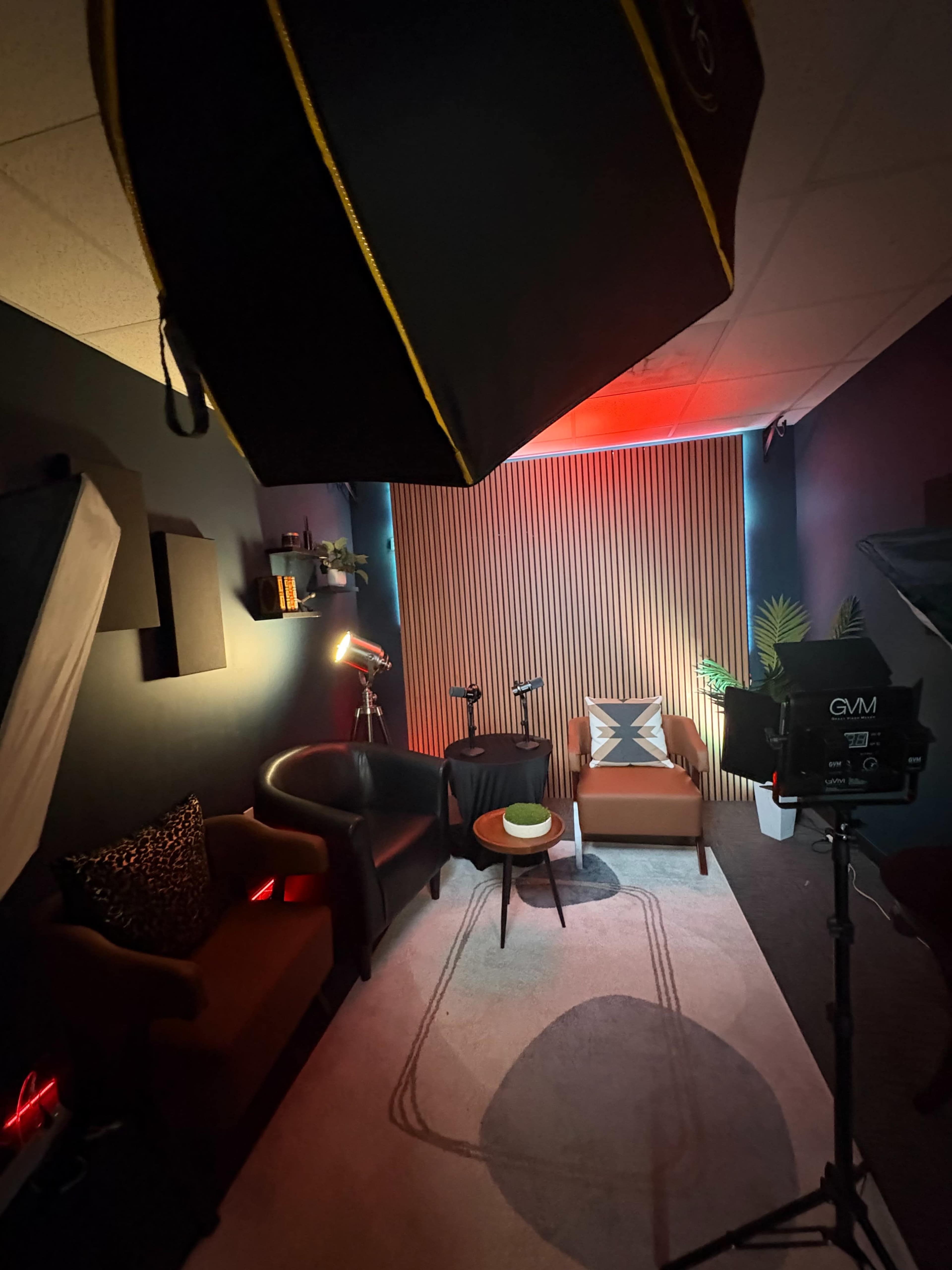 The image shows a cozy studio space with two chairs, a small table, and accent lighting against a wooden-paneled backdrop.