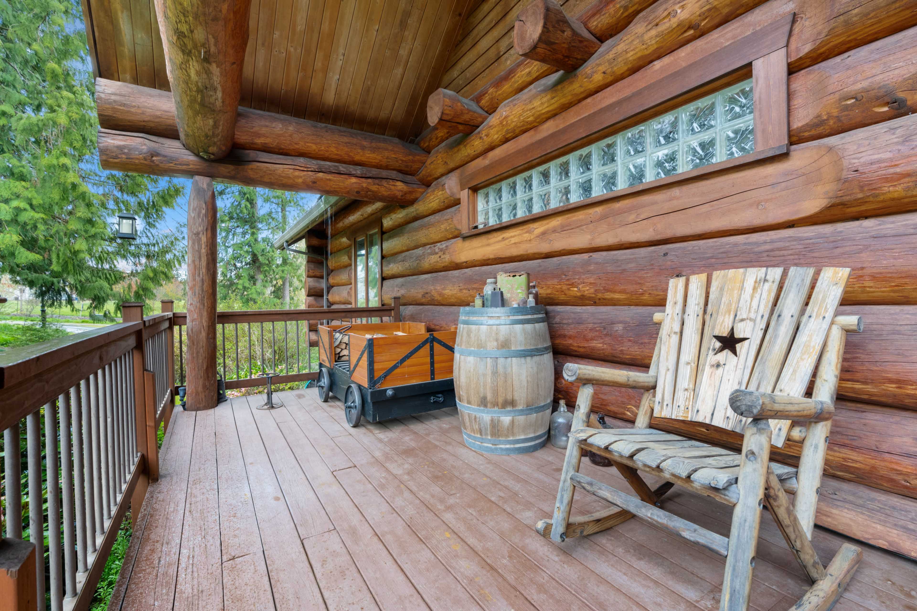 The image shows a wooden porch of a log cabin, featuring a rustic chair, a barrel, and a cart in a natural setting.