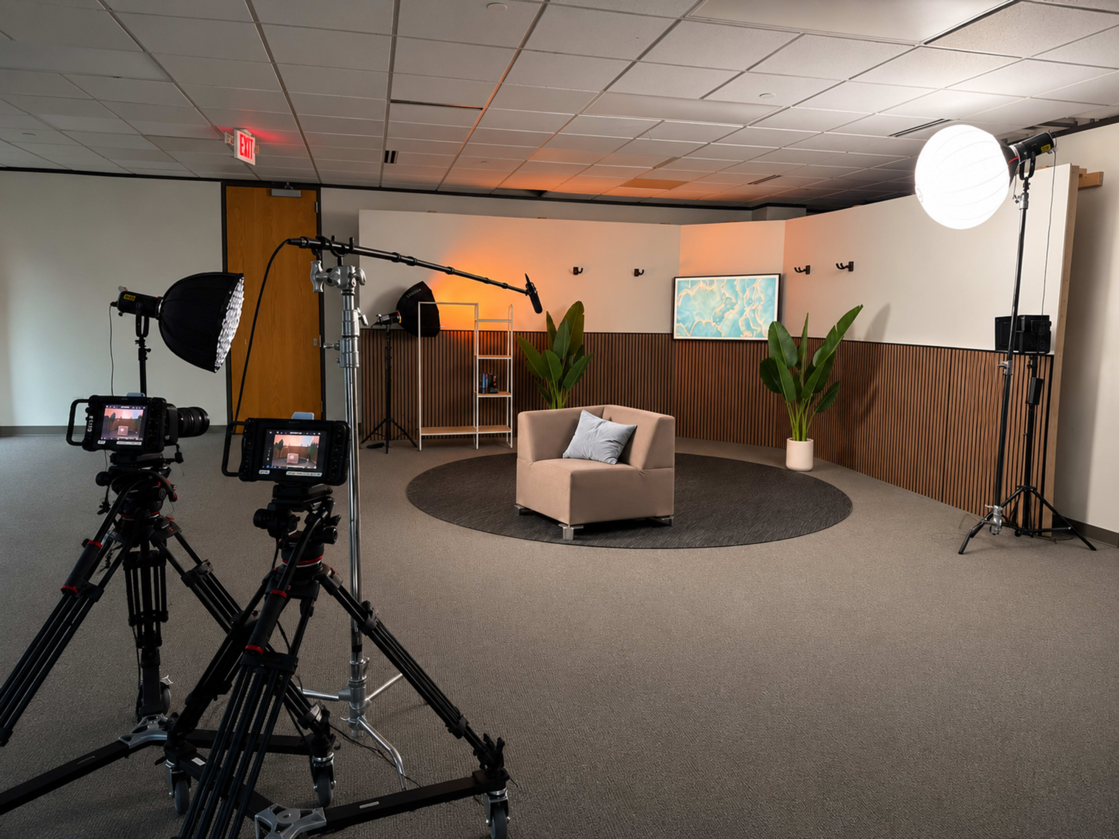 The image features a film or photography studio equipped with multiple cameras, lighting equipment, a beige armchair, and decorative plants, creating a setup for a production shoot.