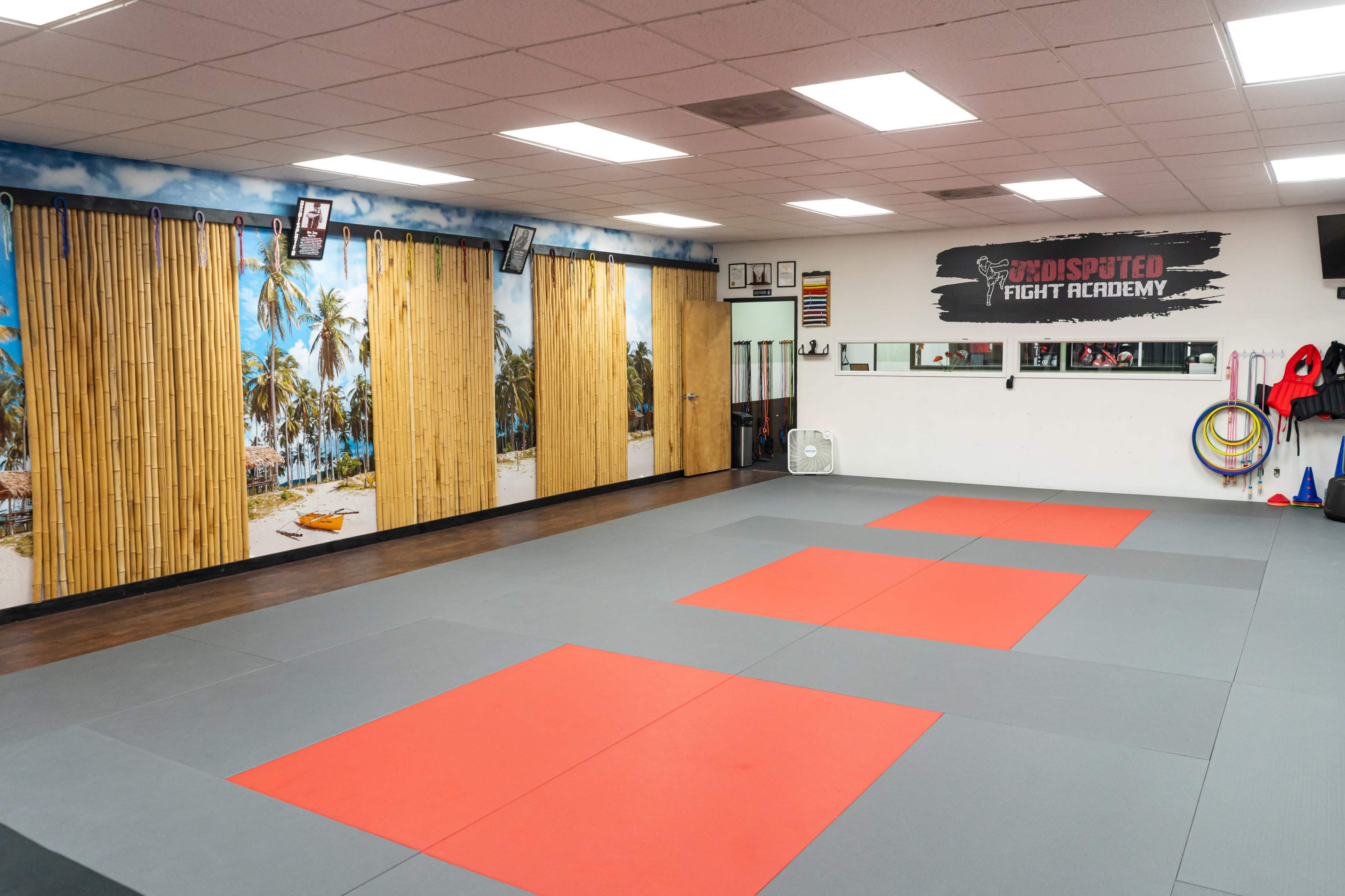 The image shows an empty martial arts training room with gray and red mats, bamboo wall decorations, and a tropical beach mural in the background.