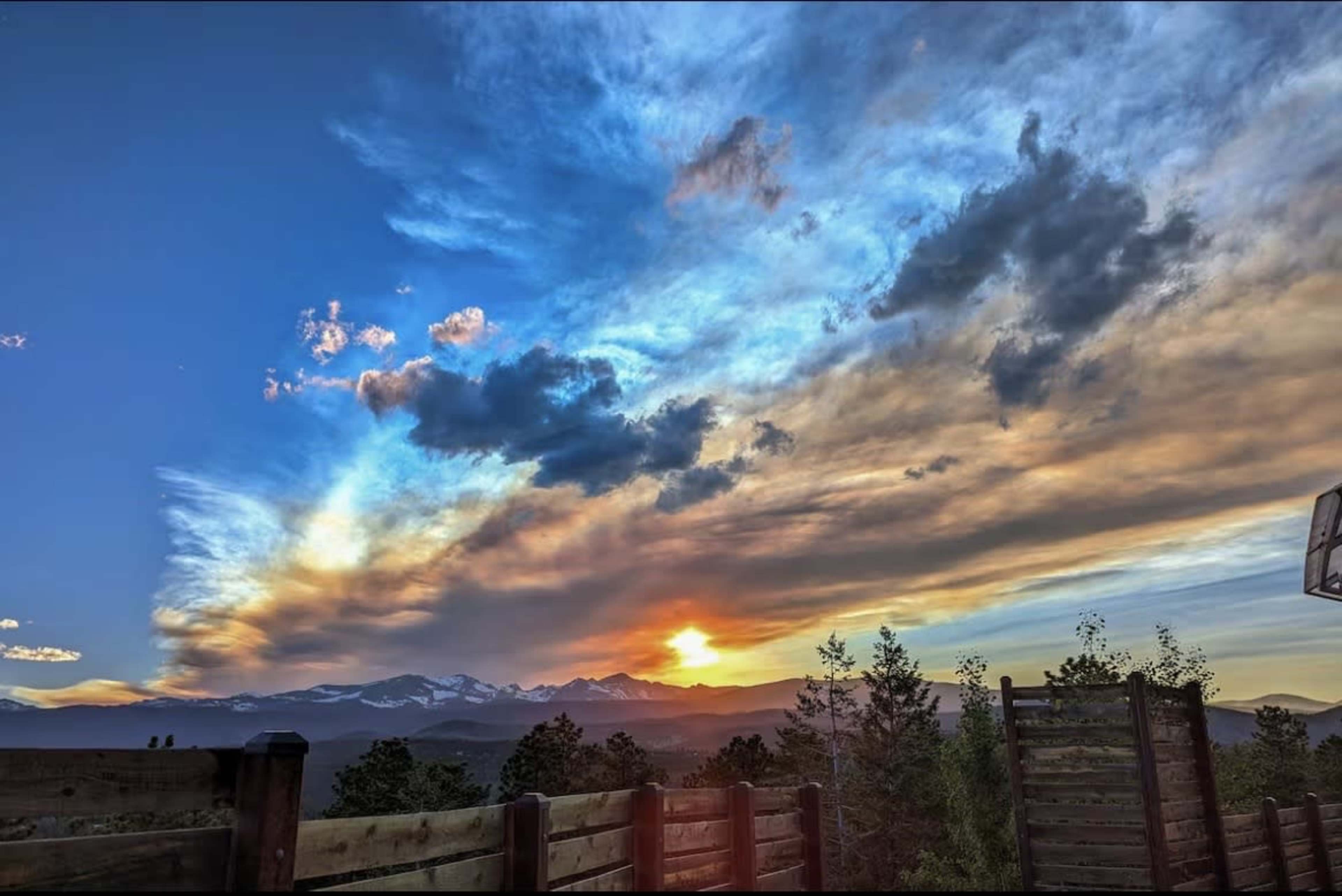 The image shows a vibrant sunset over mountains with clouds in a blue sky, framed by trees and a wooden fence in the foreground.