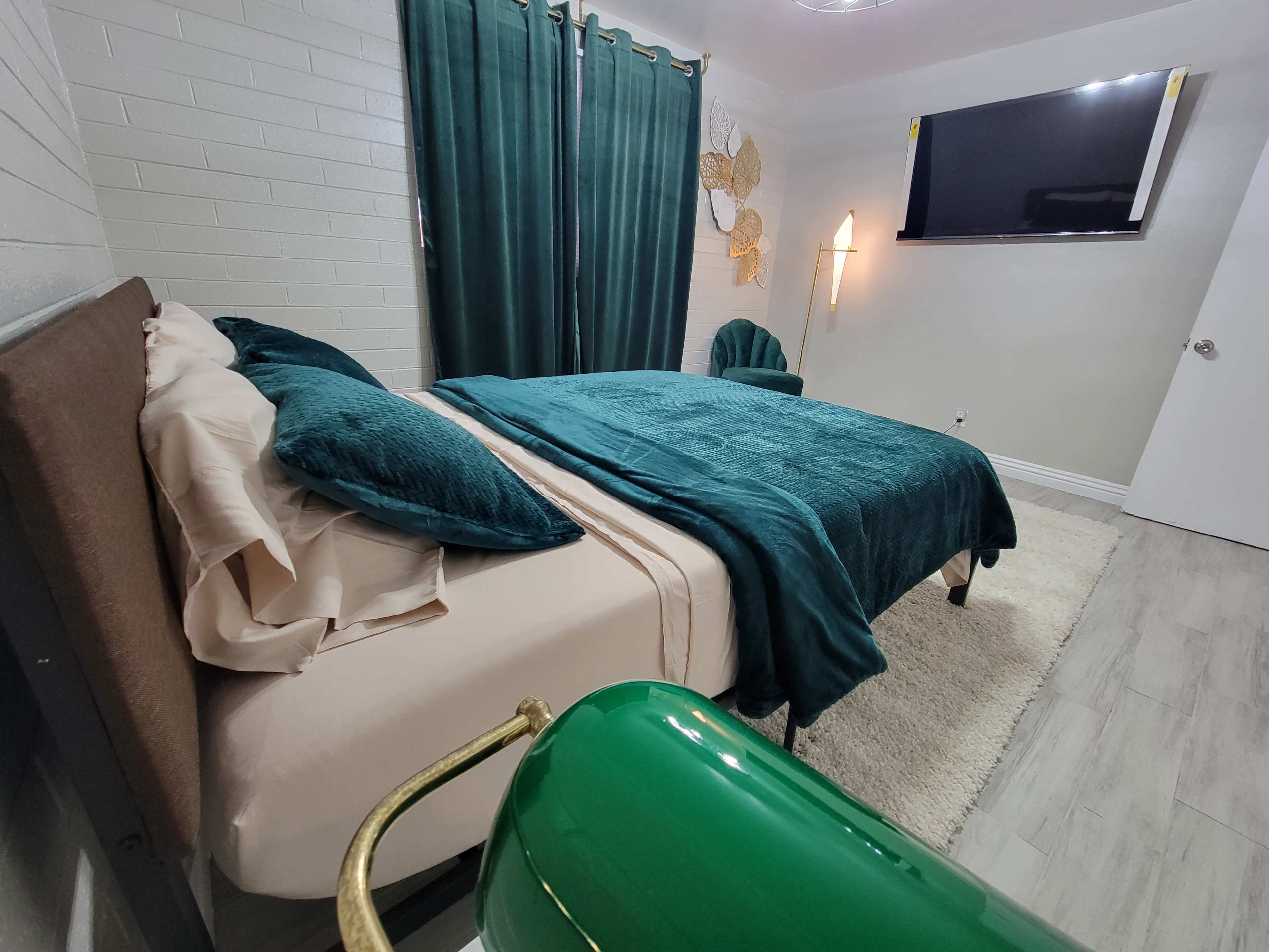 The image shows a neatly arranged bedroom featuring a green bedspread, matching curtains, and a wall-mounted television.