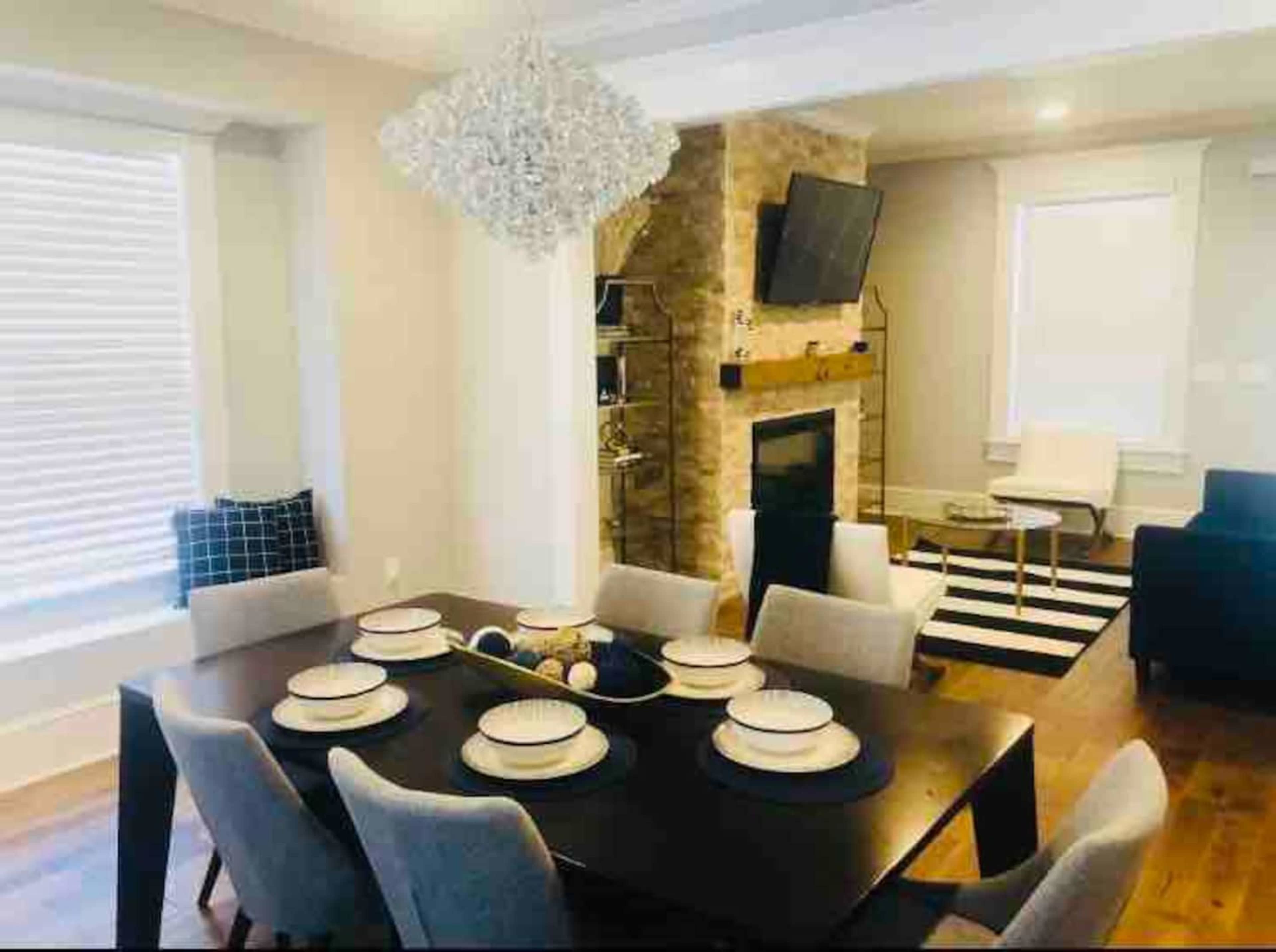 A modern dining room with a dark table set for eight, a stone fireplace, and a large chandelier above.