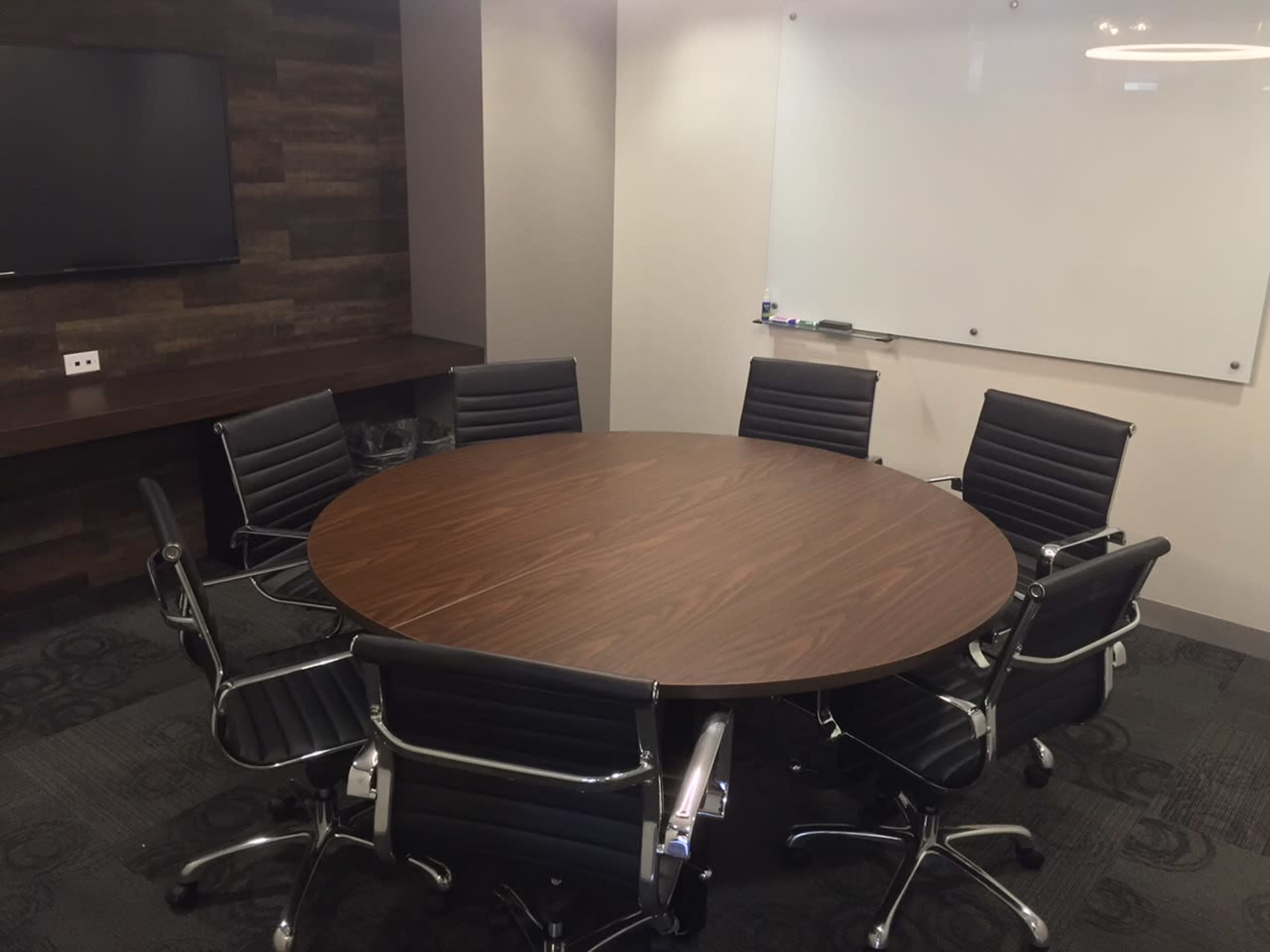 A round wooden conference table surrounded by eight black office chairs in a well-lit meeting room.