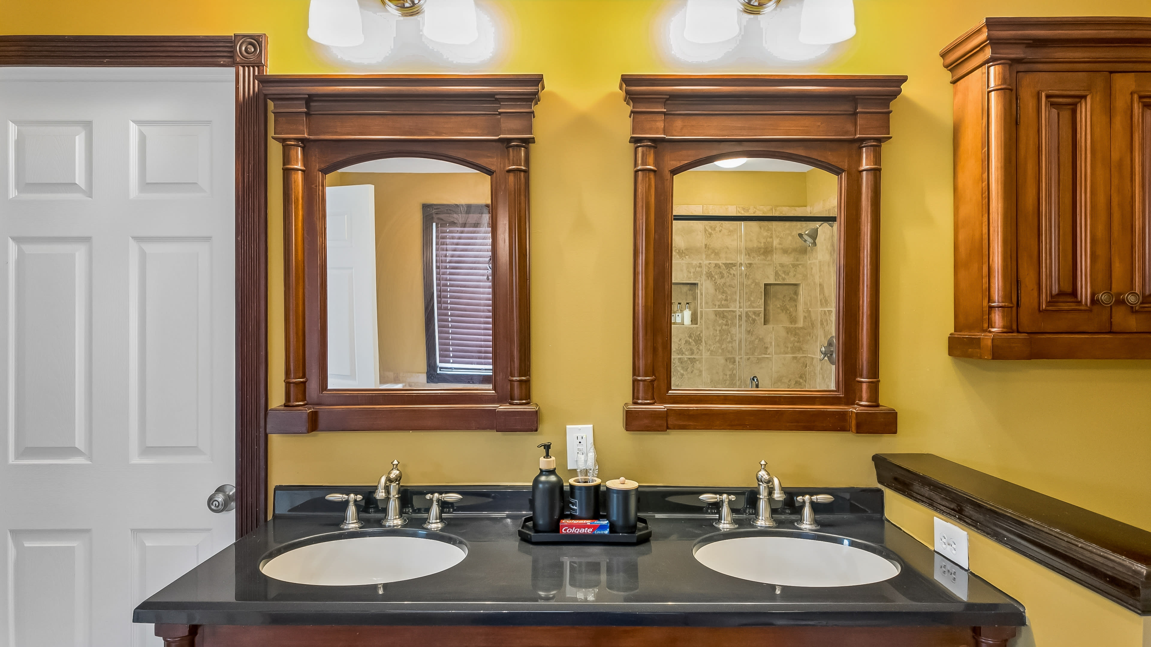 The image shows a bathroom countertop with two sinks, framed mirrors above them, and wooden cabinetry against a yellow wall.