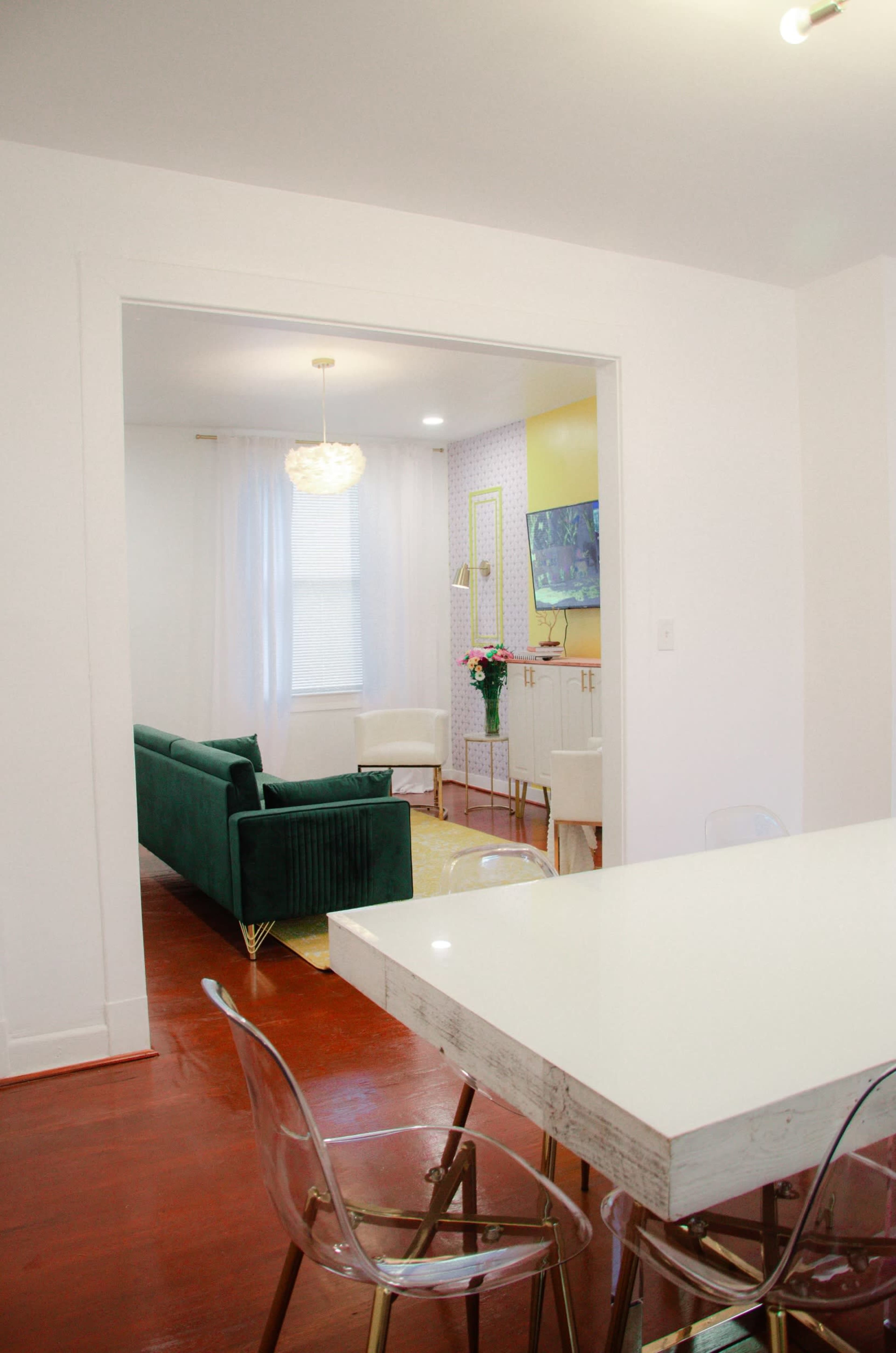 A dining area with a white table and clear chairs opens into a living room featuring a green couch, white curtains, and modern decor.