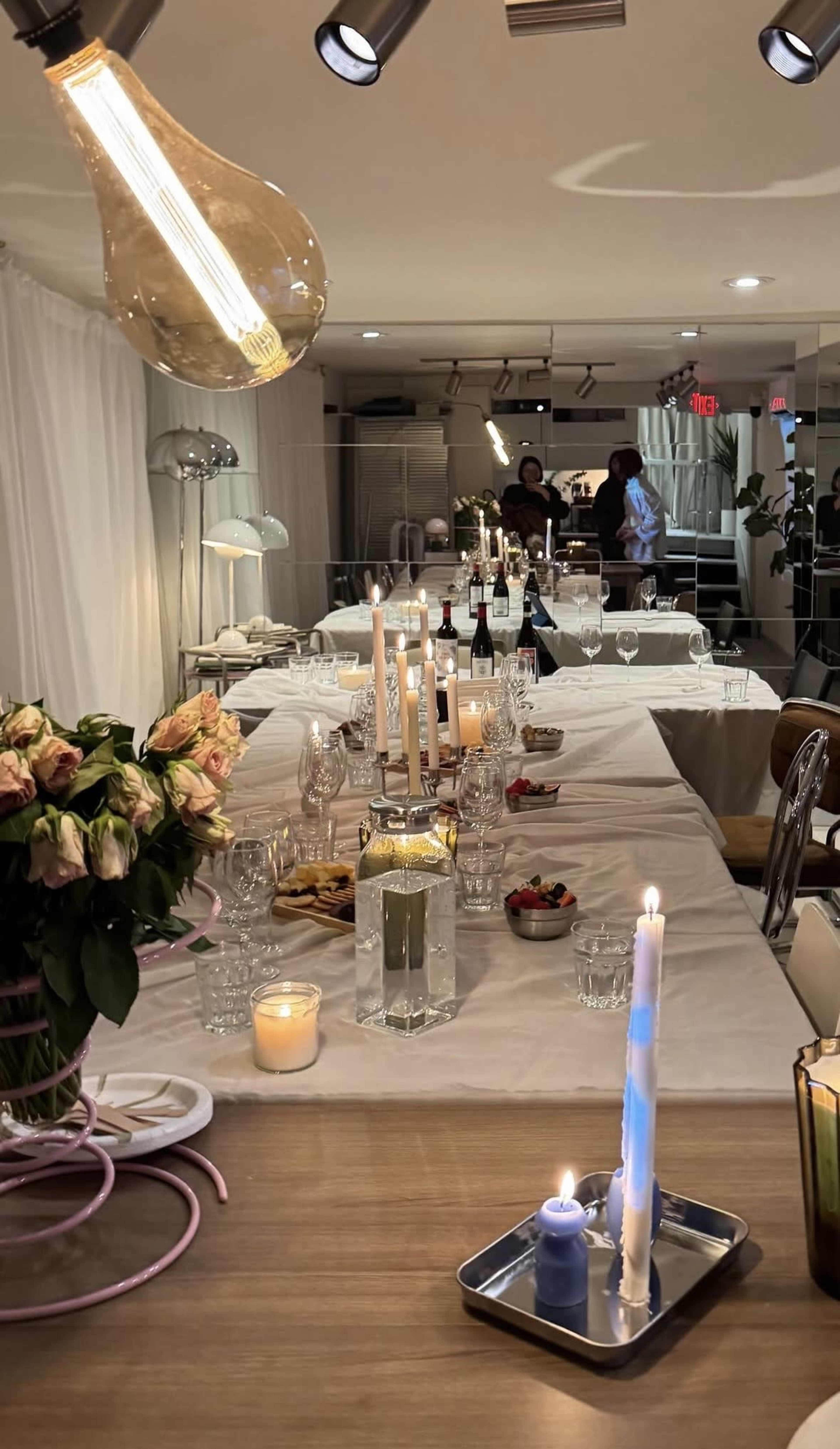 A long dining table is set with candles, wine bottles, and plates of food, with a bouquet of flowers at one end and soft lighting throughout the room.