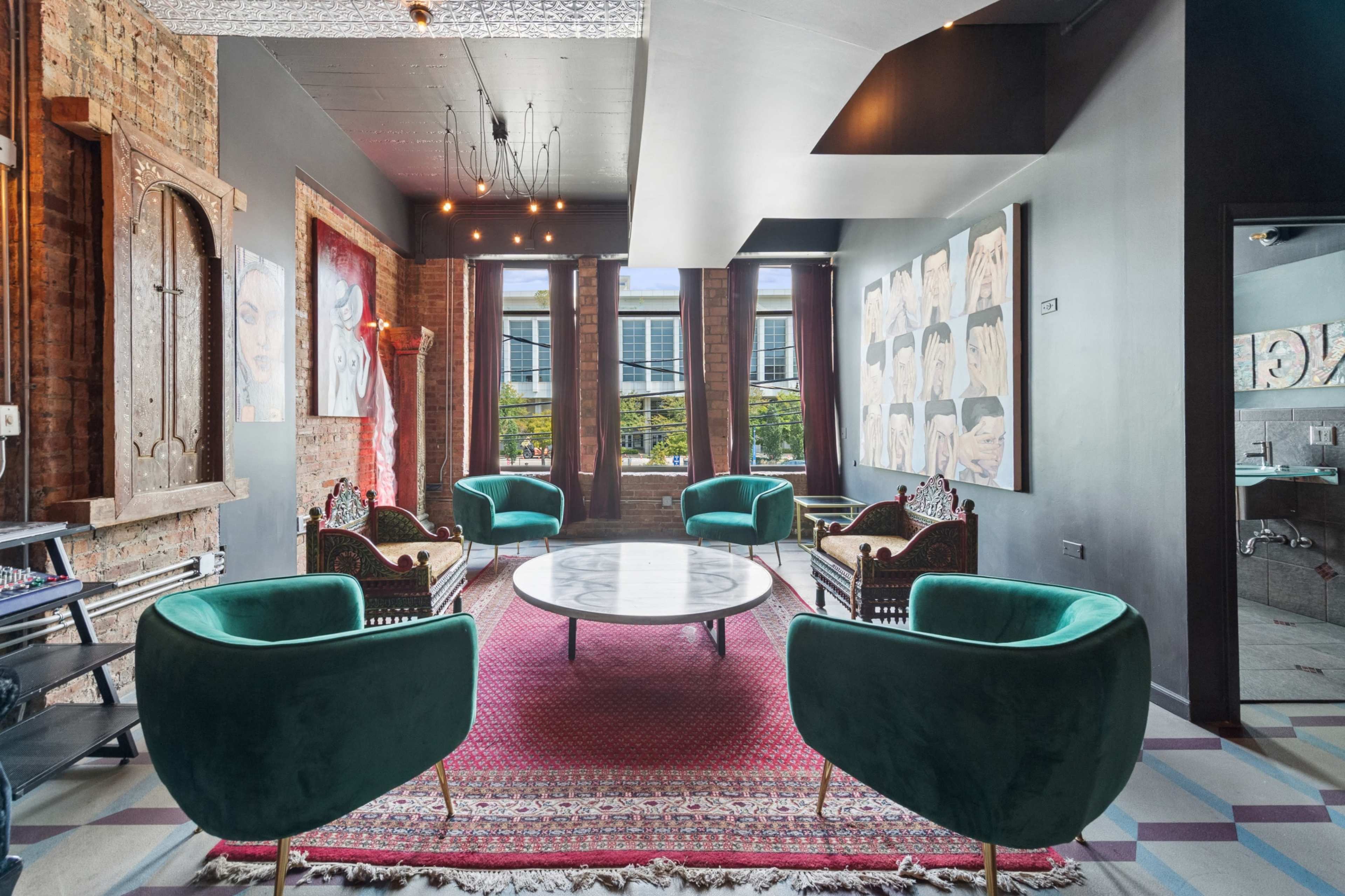 The image shows a stylish lounge area with modern green chairs, a round marble table, and artwork displayed on the walls, illuminated by natural light from large windows.