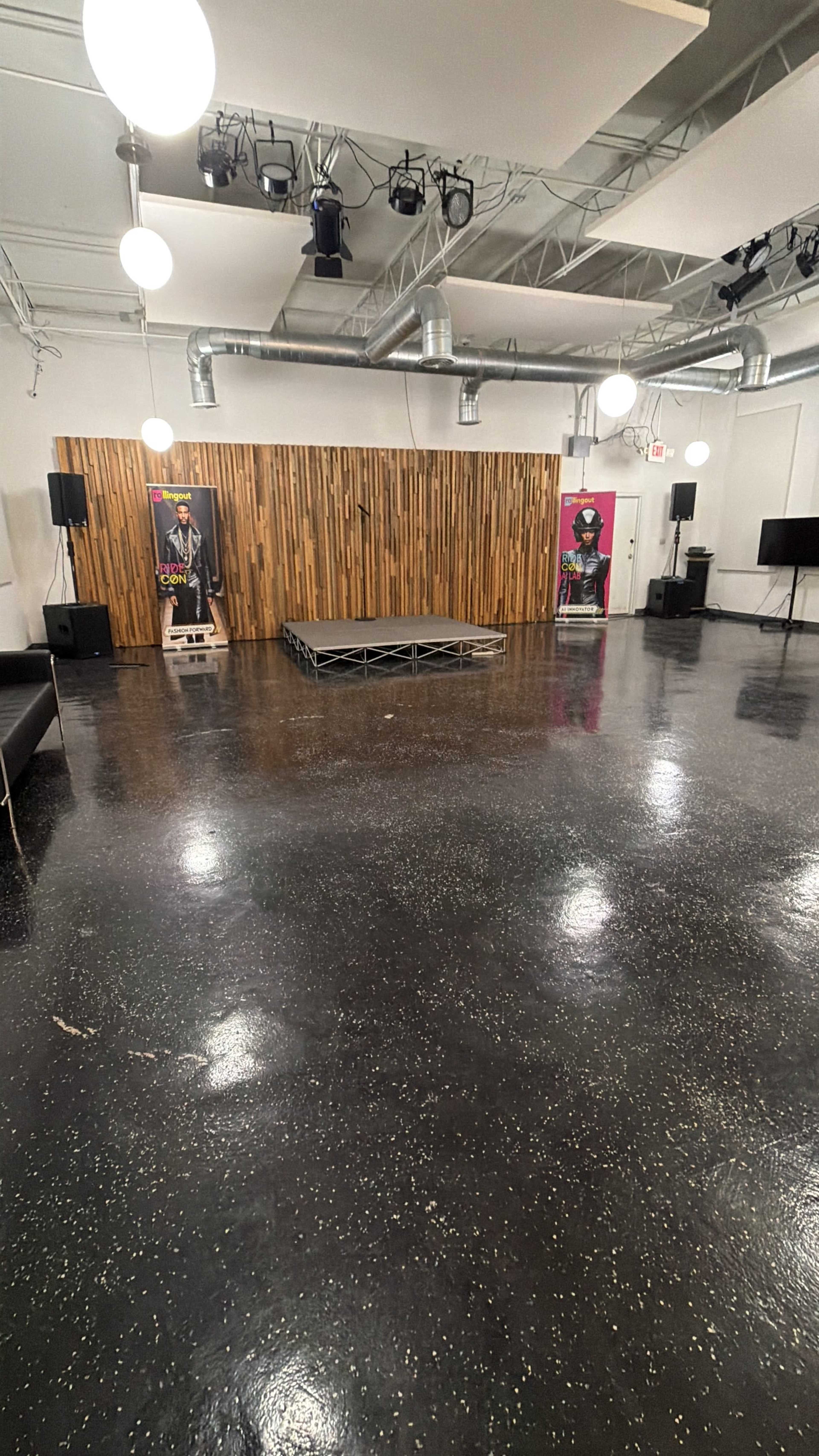 The image shows a spacious room with a wooden wall at one end, a small stage in the center, and various lighting fixtures hanging from the ceiling.